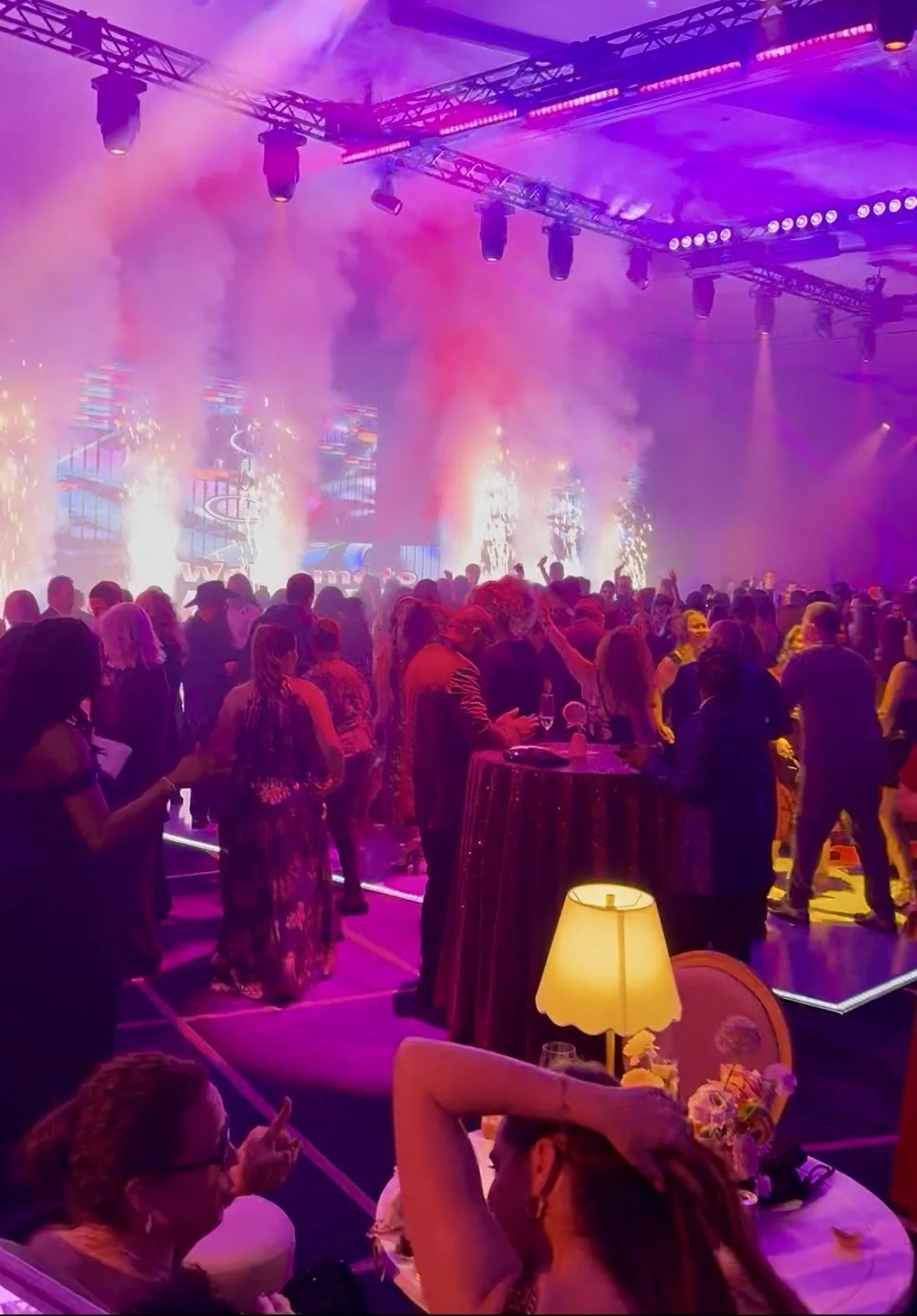 People dancing and socializing at a vibrant indoor event with colorful lighting, smoke effects, and live performance on stage.