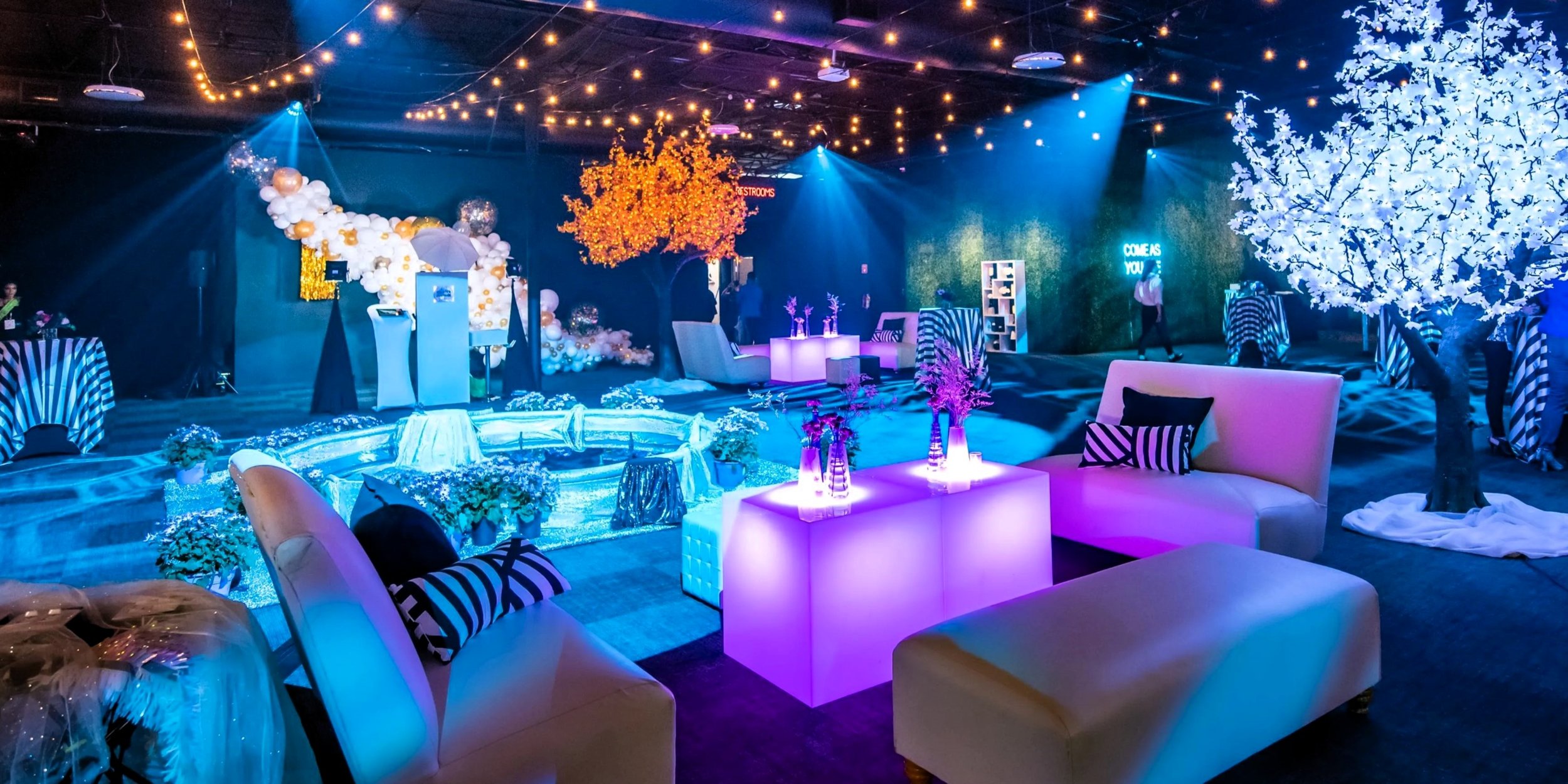 Indoor event space decorated with colorful lighting, fairy lights, artificial trees, and floral arrangements. Seating includes white sofas and striped cushions, with illuminated tables and a balloon display in the background.