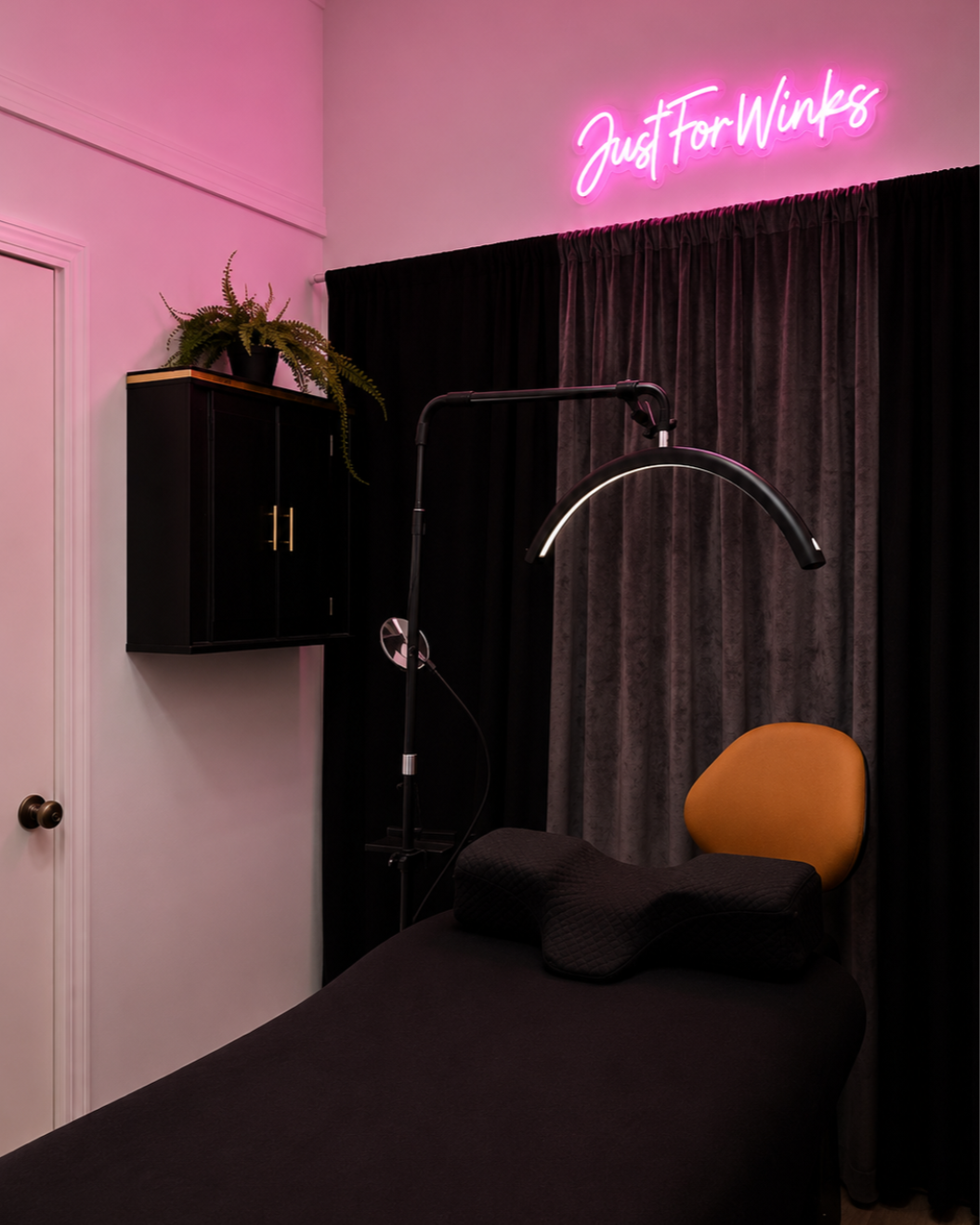 A massage or therapy room with a massage table with a black cover, a black pillow, and a yellow headrest. There's a dark curtain, a black wall cabinet with a potted fern, a black floor lamp, and a pink neon sign that reads 'Just For Winks'.