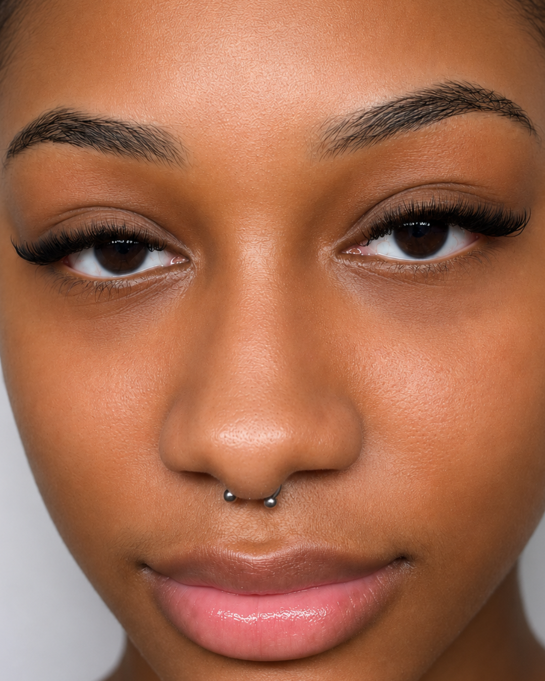 Close-up of a woman with dark skin, brown eyes, long eyelashes, and a septum piercing.