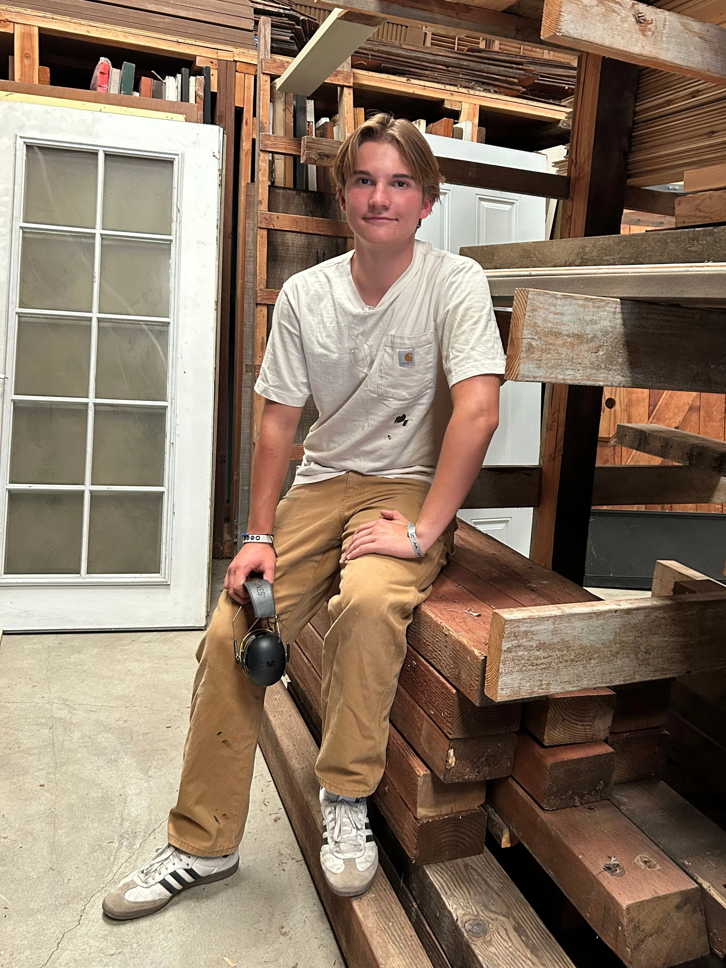 A young man sitting on a wooden staircase in a workshop or lumber yard. He is wearing a white t-shirt, tan pants, and sneakers, holding headphones in his right hand. There are wooden planks and building materials around him, with a white door and shelving in the background.