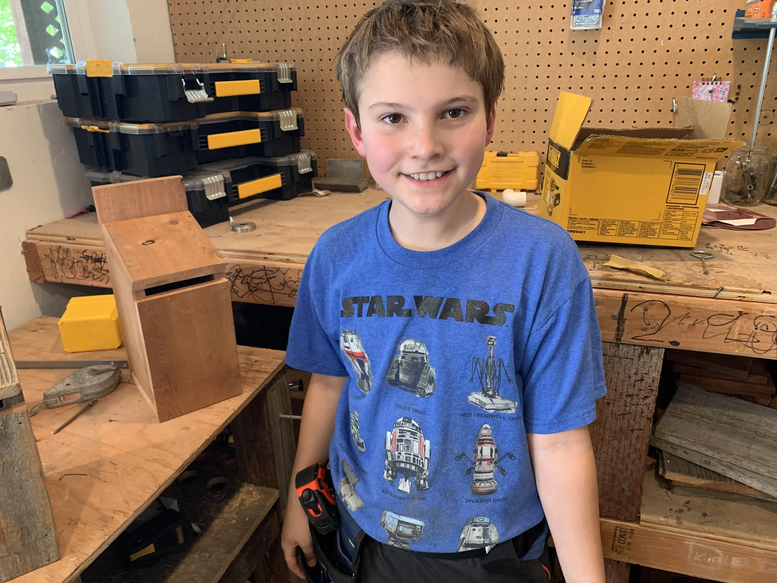 A young boy with brown hair and a wide smile stands in a workshop, wearing a blue Star Wars T-shirt. Behind him is a pegboard wall, a window, and a workbench with tools and storage boxes.