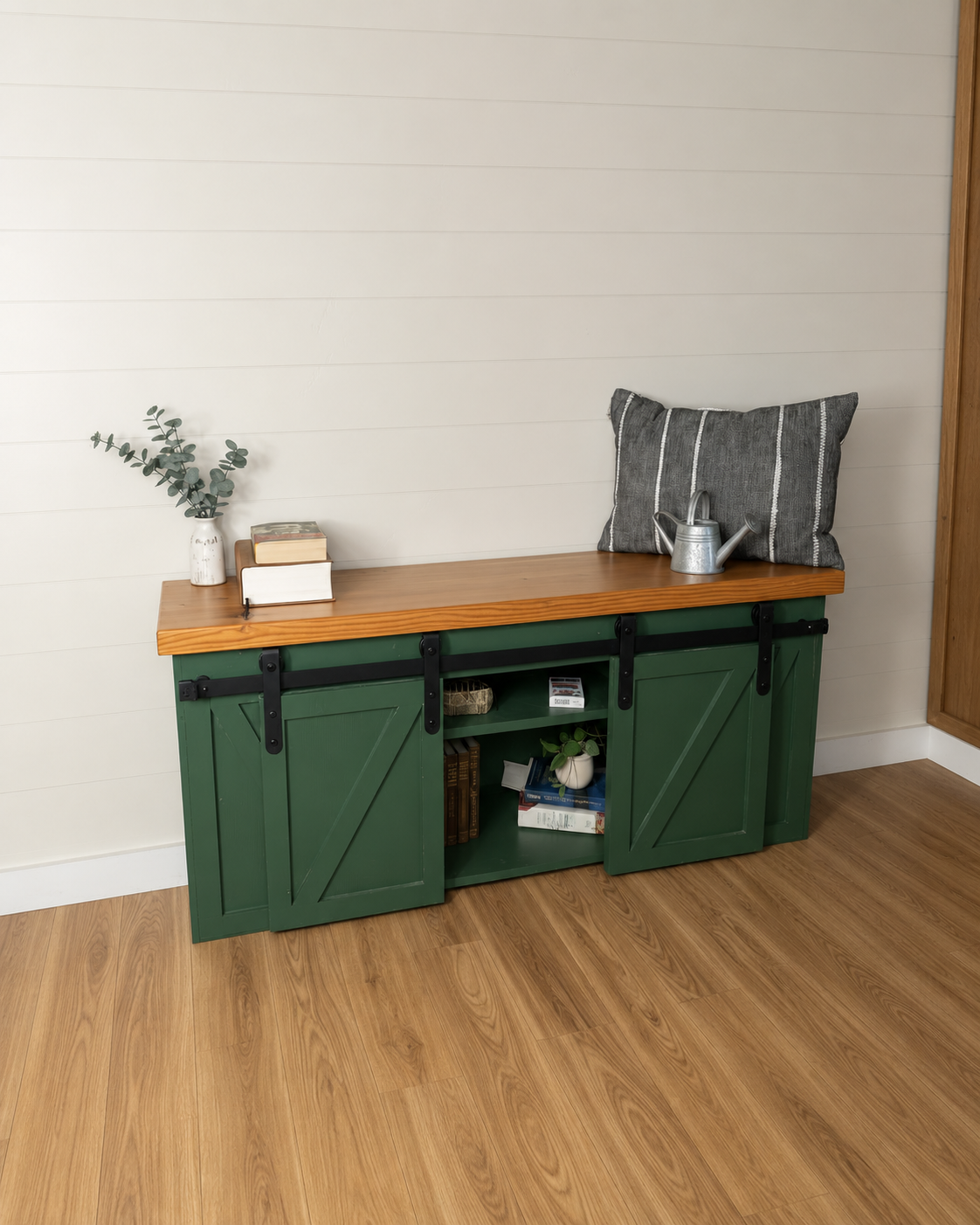 A green wooden cabinet with sliding barn doors, topped with a wooden surface. On top, there is a white vase with eucalyptus, a small stack of two books, a gray striped pillow, and a small silver watering can. The lower shelves of the cabinet contain books and a small potted plant. The room has light-colored wooden flooring and a white wall.