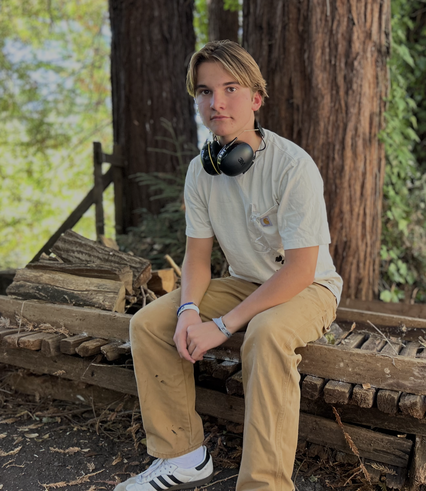 Young man sitting on a wooden log wearing a white T-shirt, khaki pants, white sneakers, and black headphones in a forested area.