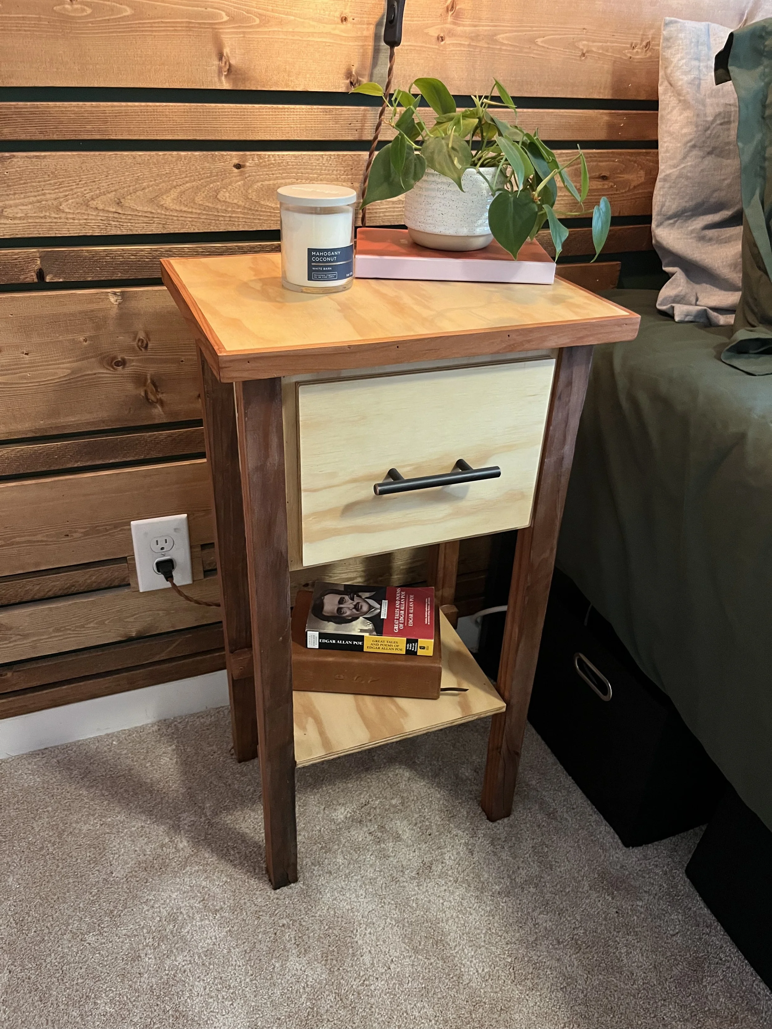 A wooden side table with a light-colored drawer and a black handle, standing on a beige carpeted floor. On top of the table, there is a potted plant with green leaves, a pink book, and a white candle in a glass jar labeled 'Mahogany Coconut.' There is a wooden wall in the background, an electrical outlet on the wall, and a bed with dark green bedding and a gray pillow to the right.