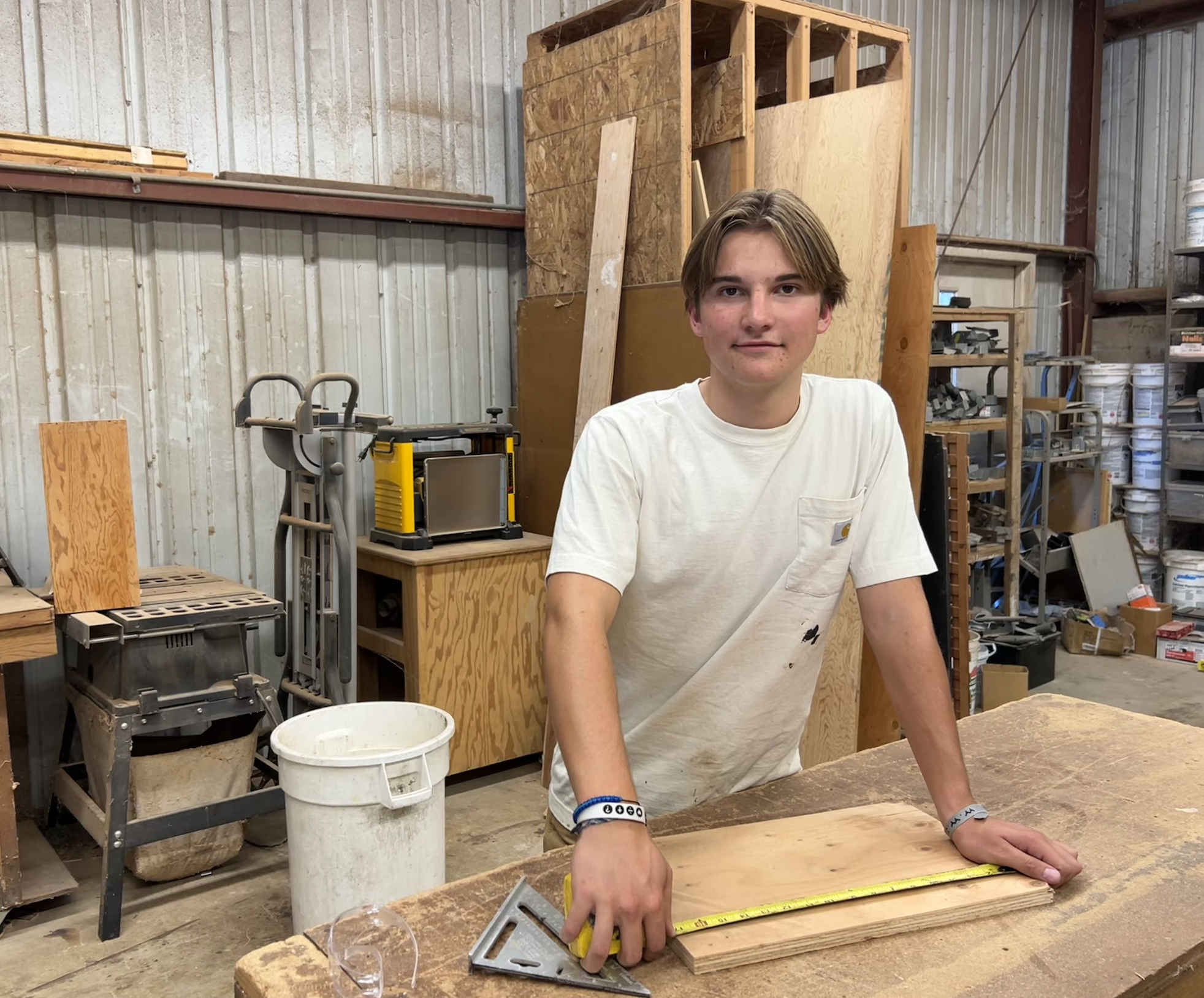A young man with light brown hair working in a woodworking shop, measuring a piece of wood with a yellow tape measure, surrounded by various tools, wood, and supplies.