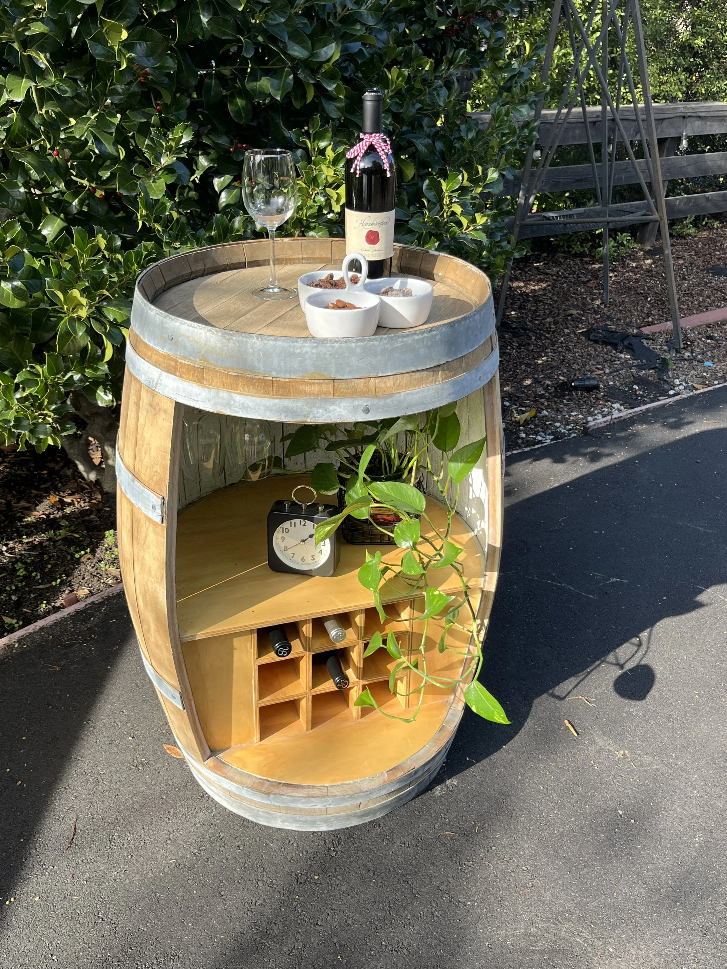A whiskey barrel wine rack with a wine bottle, an empty wine glass, and three small bowls of snacks on top, next to green bushes and a gray garden structure.