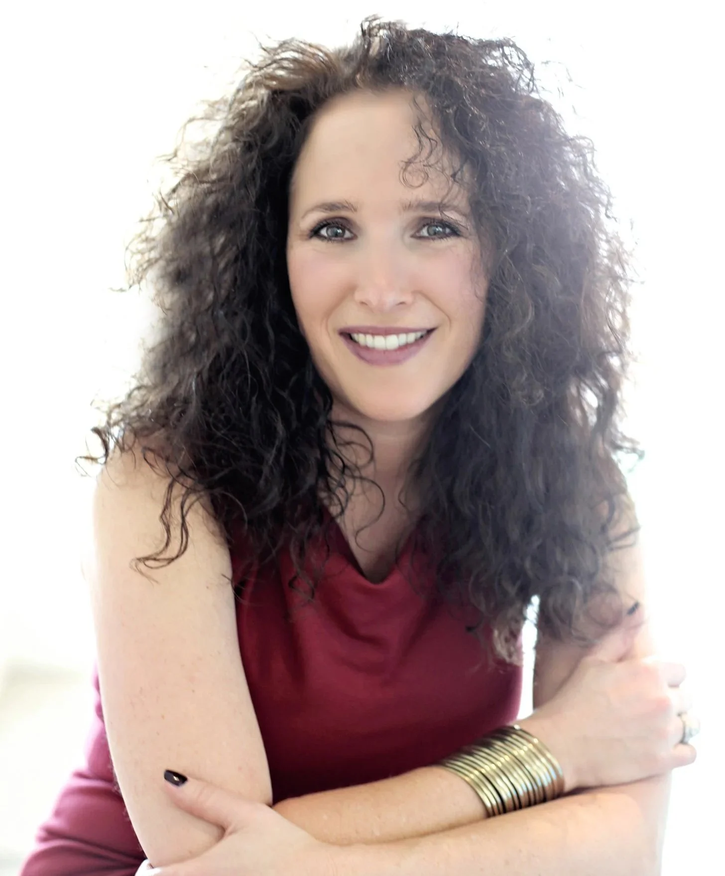 Author Aimee Cohen, a woman with curly dark hair smiling, her arms crossed in front of her, wearing a red dress and gold bangles.