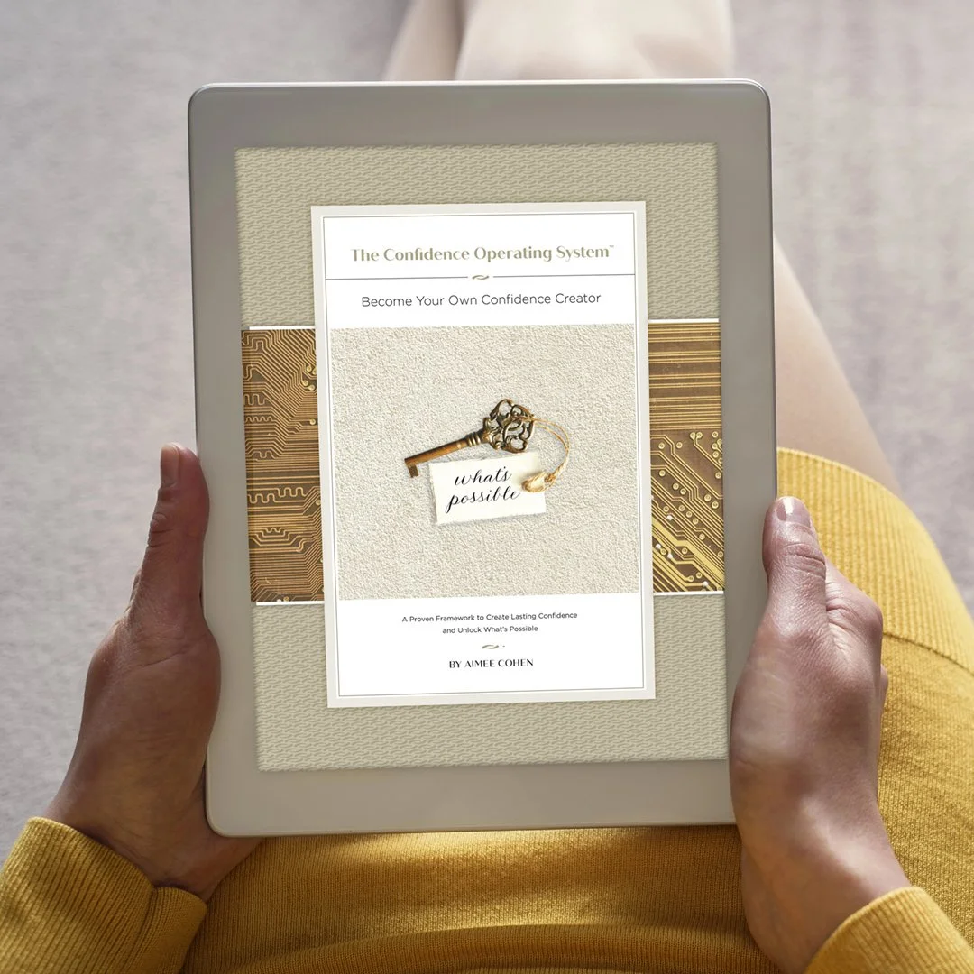 A person wearing a yellow long sleeve shirt sitting on a couch or chair, holding a tablet displaying a book cover titled 'The Confidence Operating System' by Aimee Cohen, with a picture of a key and a note that says 'what's possible' on the cover.