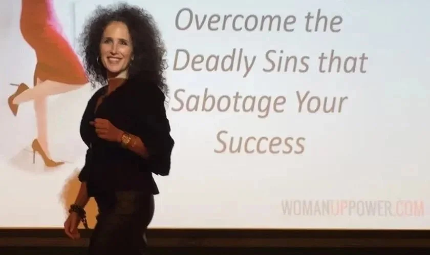 Author Aimee Cohen, a woman with curly hair smiling, standing in front of a presentation slide that reads 'Overcome the Deadly Sins that Sabotage Your Success' with WOMAN UP! in the corner.