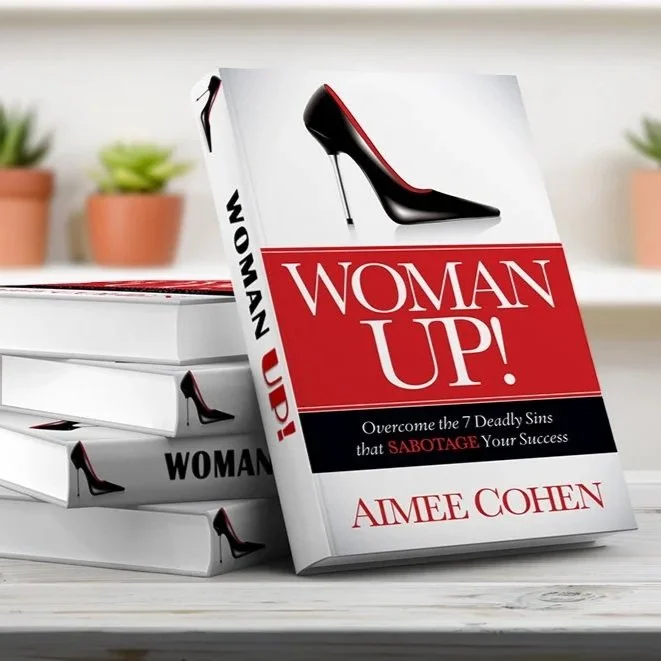 Stack of books titled 'Woman Up!' with a high-heeled shoe graphic, on a white surface with potted plants in the background.