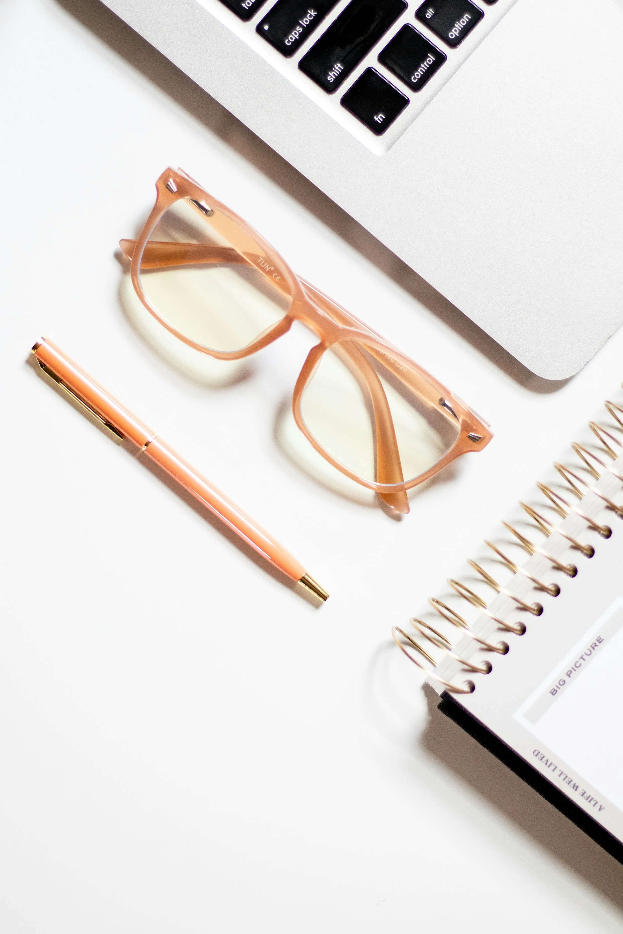 Close-up of a workspace with a laptop keyboard, pink eyeglasses, a pink pen, a spiral notebook, and a partially visible book on white surface.