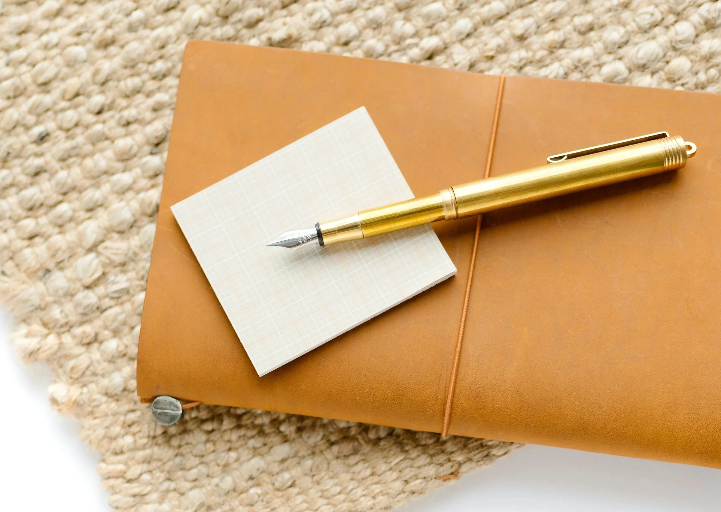 A gold pen resting on a small notepad with graph paper pages, placed on a brown leather-bound notebook or journal, on a textured beige carpet.