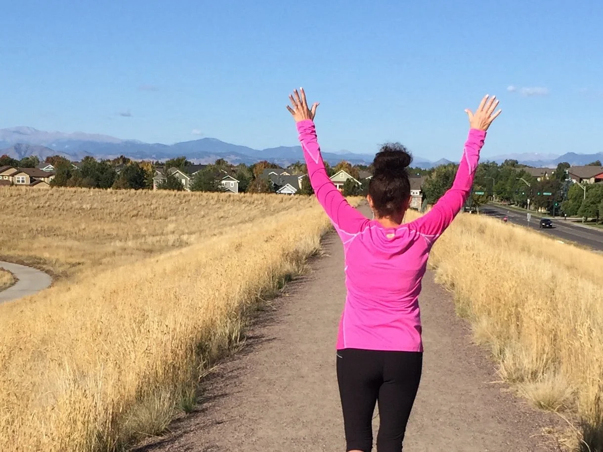 Aimee Cohen, confidence coach, a woman in a pink long-sleeve shirt and black leggings walking away from the viewer on a dirt path with her arms raised in the air, surrounded by tall yellow grass with houses and mountains in the background.
