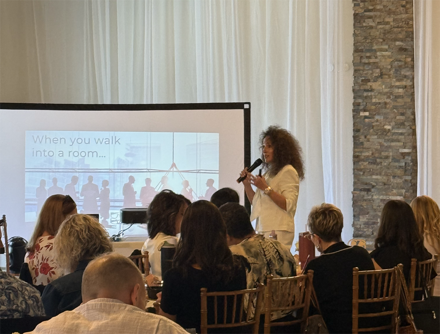 Aimee Cohen, confidence creator and speaker, a woman with curly hair giving a presentation on a stage in front of an audience, with a screen displaying a slide that reads 'When you walk into a room...' and silhouettes of people in the background.