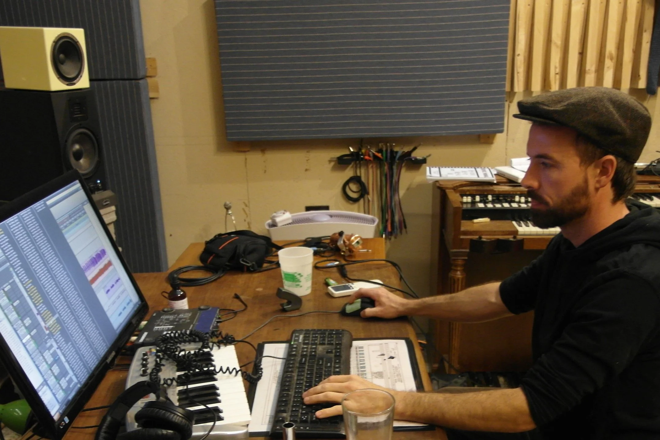A man wearing a brown flat cap working in a recording studio. He is seated at a wooden desk with a computer, mixing console, and various audio equipment. There are large speakers on the wall behind him, a pair of glasses, a cup, and a smartphone on the desk. The studio has soundproofing panels on the walls.