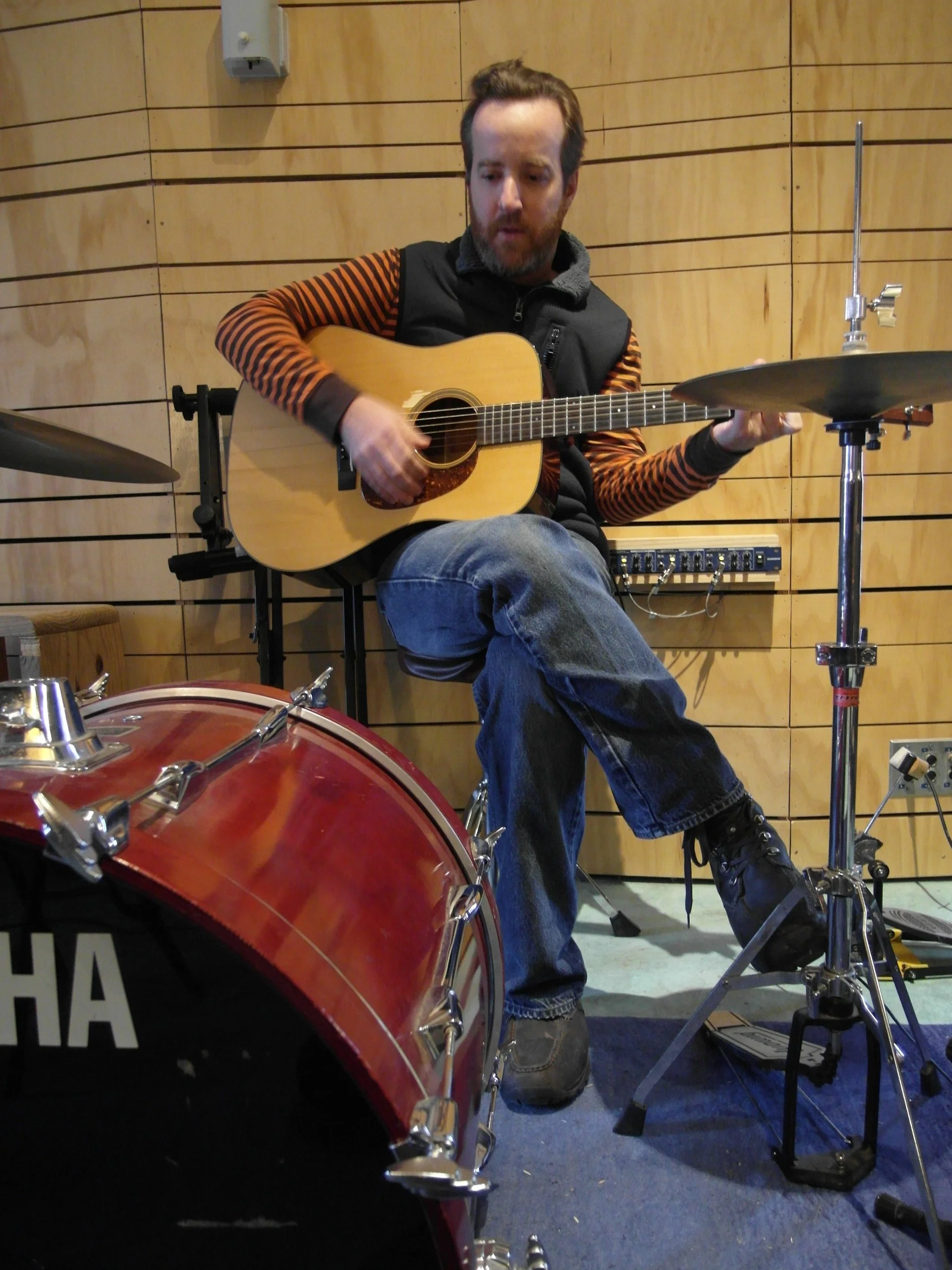 A man with a beard and wearing a black vest and striped orange and black long sleeve shirt playing an acoustic guitar in a room with wooden panel walls, next to a drum set and music equipment.