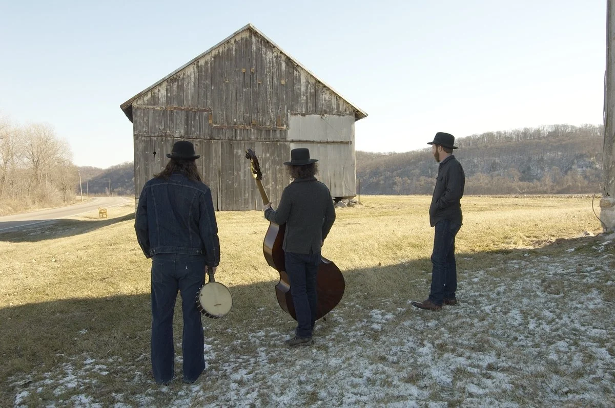 Three men wearing black hats and dark clothing standing on a grassy area near an old weathered barn in a rural landscape, with one man holding a guitar and another holding a tambourine.