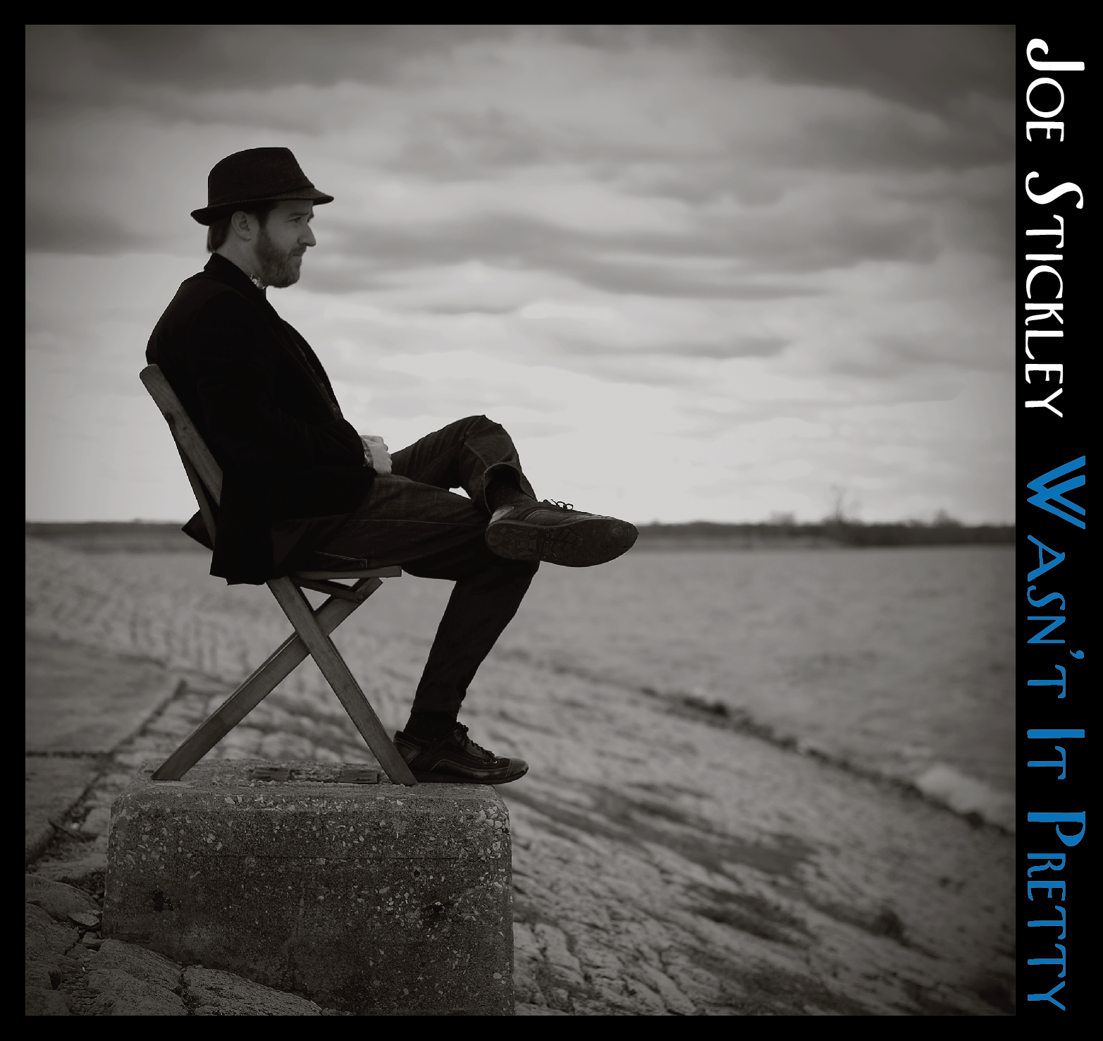 A man sitting on a folding chair near a body of water, looking out at the cloudy sky, in black and white.