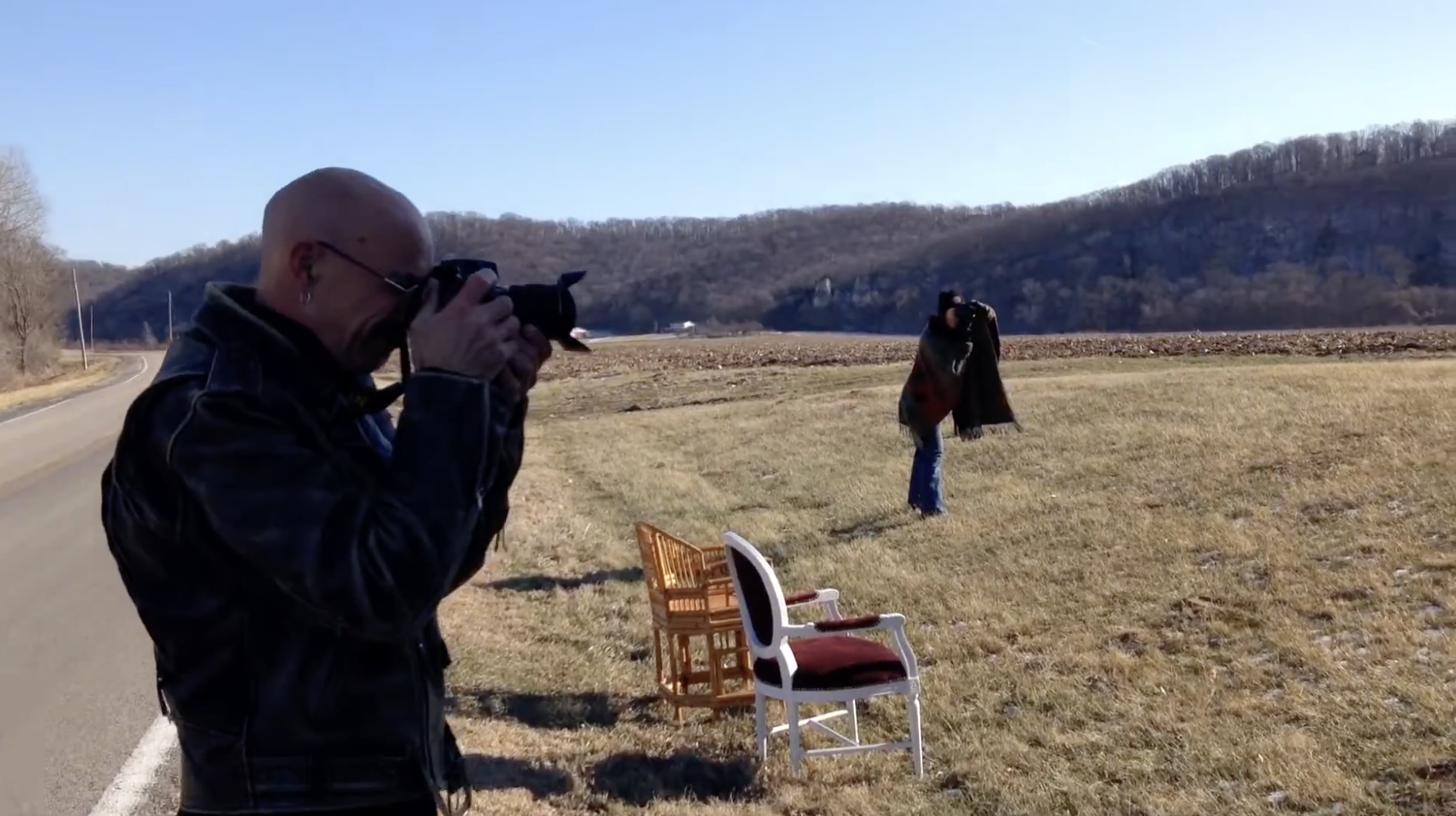 A man with a bald head, glasses, and earrings is taking a photograph outdoors with a camera. In the background, a woman is standing on a grassy field, adjusting her hat or headwear. There are three chairs set up in the grass, and the scene is set against a backdrop of mountains and a clear blue sky.