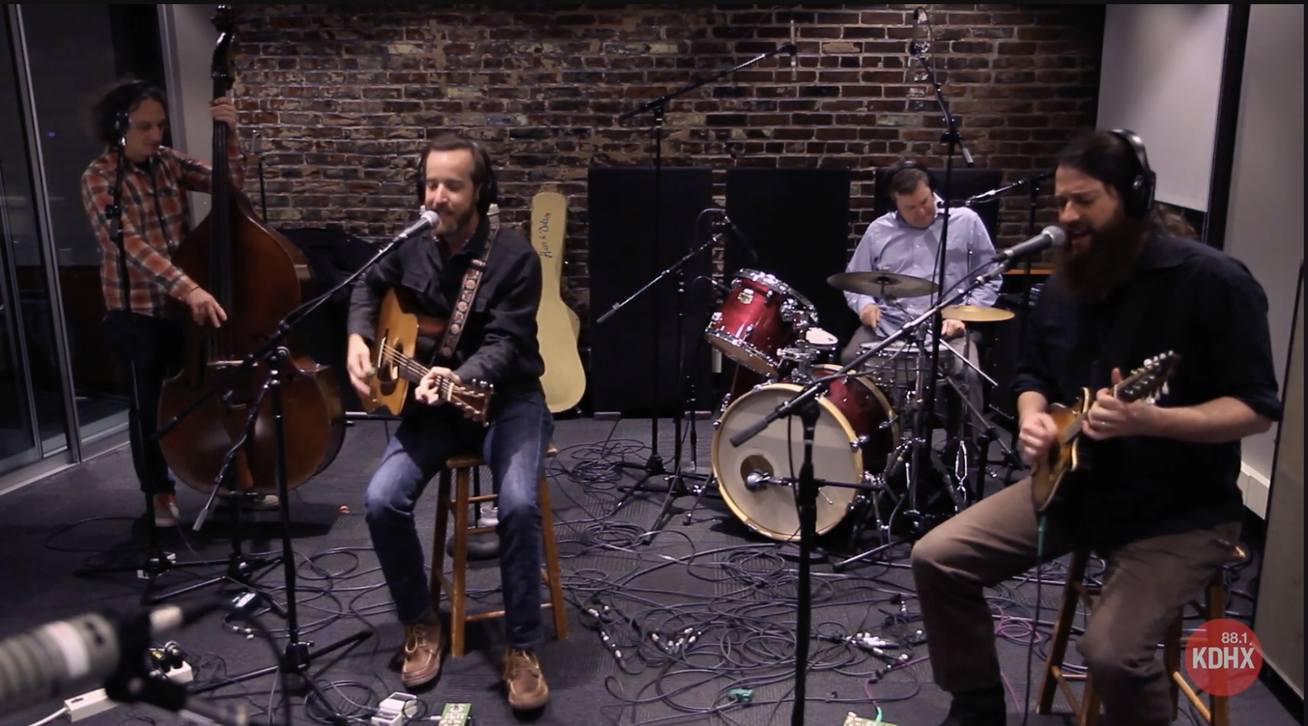 A band of four musicians performing in a studio, with a brick wall background. One is playing upright bass, one is singing and playing an acoustic guitar, one is on drums, and one is playing an electric guitar.