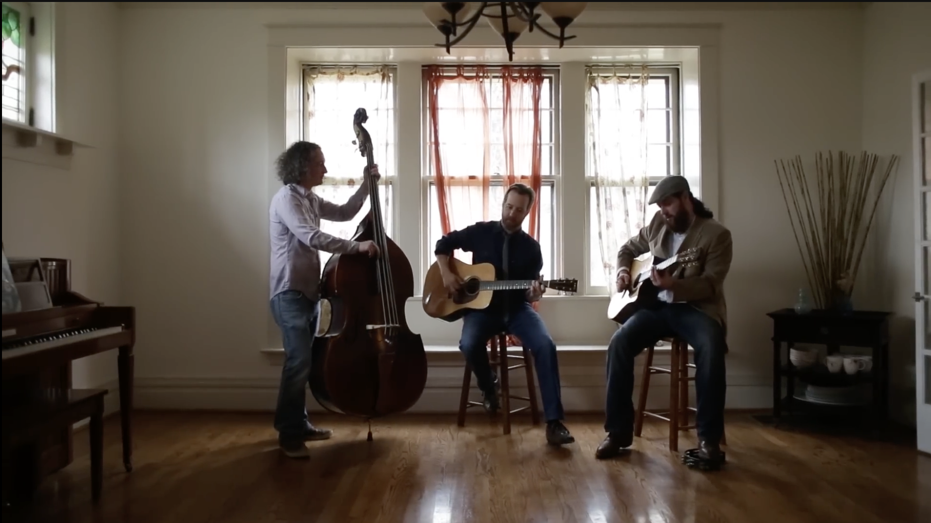 Three men playing musical instruments in a cozy room with a large window. One is on upright bass, one on acoustic guitar, and one on electric guitar. The room has a piano, a side table, and decorative vases.