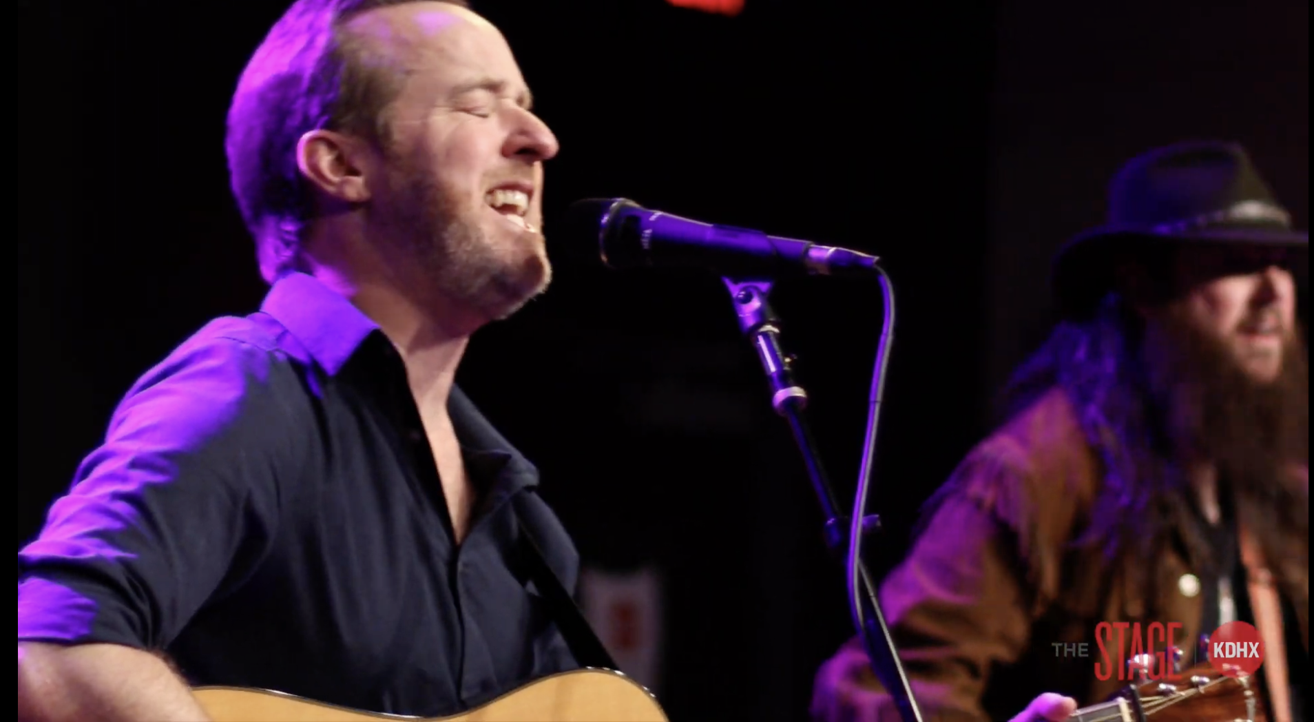 A man with short hair, beard, and black shirt, singing into a microphone while playing an acoustic guitar on stage, with another man in the background wearing a hat and glasses, also playing guitar.