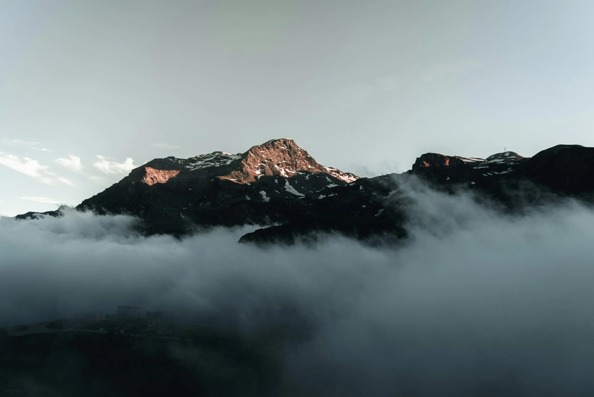 Montagnes rocheuses avec quelques neiges, entourées de nuages et une légère brume au sol, sous un ciel clair.