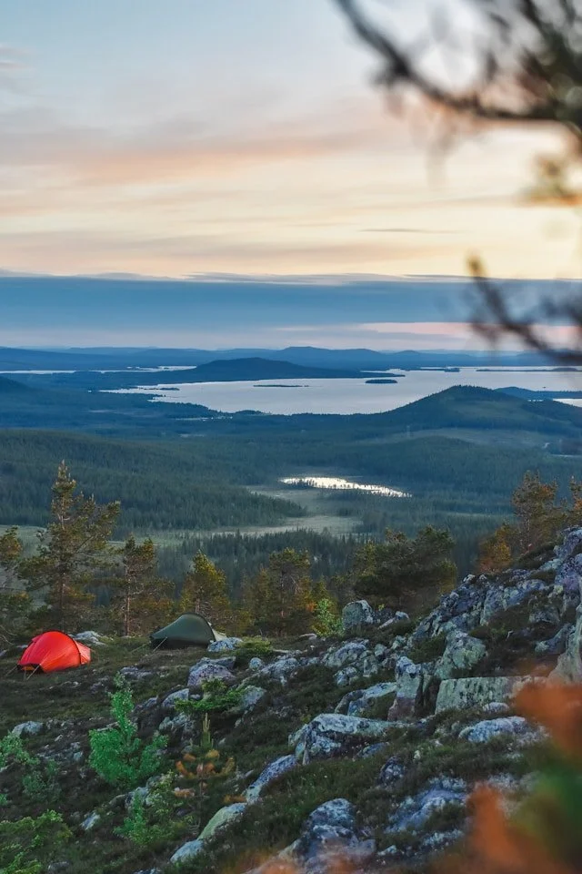 Tentes colorées sur un terrain rocheux au sommet d'une montagne, avec une vue sur des lacs et des forêts au coucher du soleil.