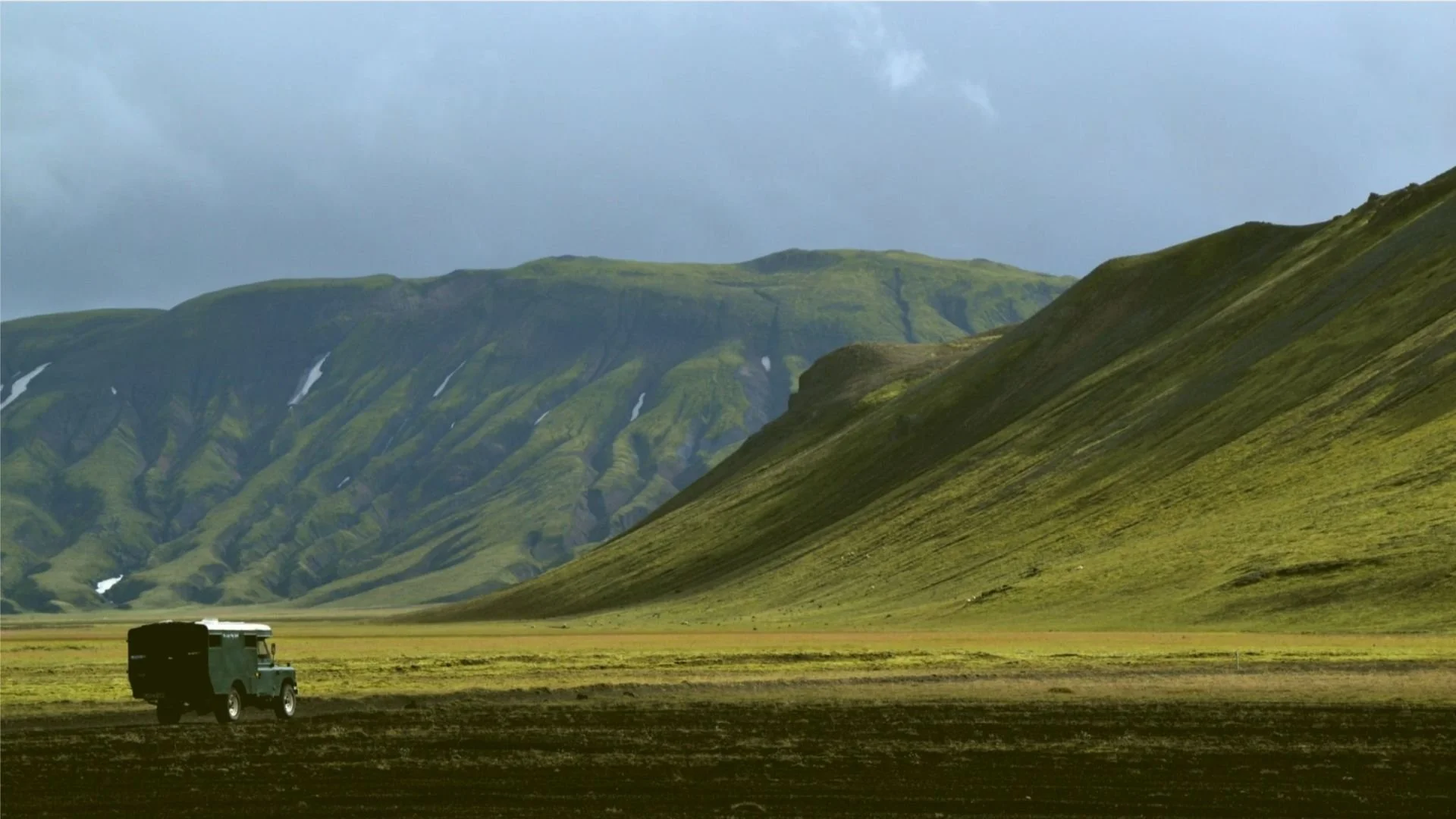 Paysage de montagne vertes avec un ciel nuageux et un véhicule tout-terrain noir dans un champ.