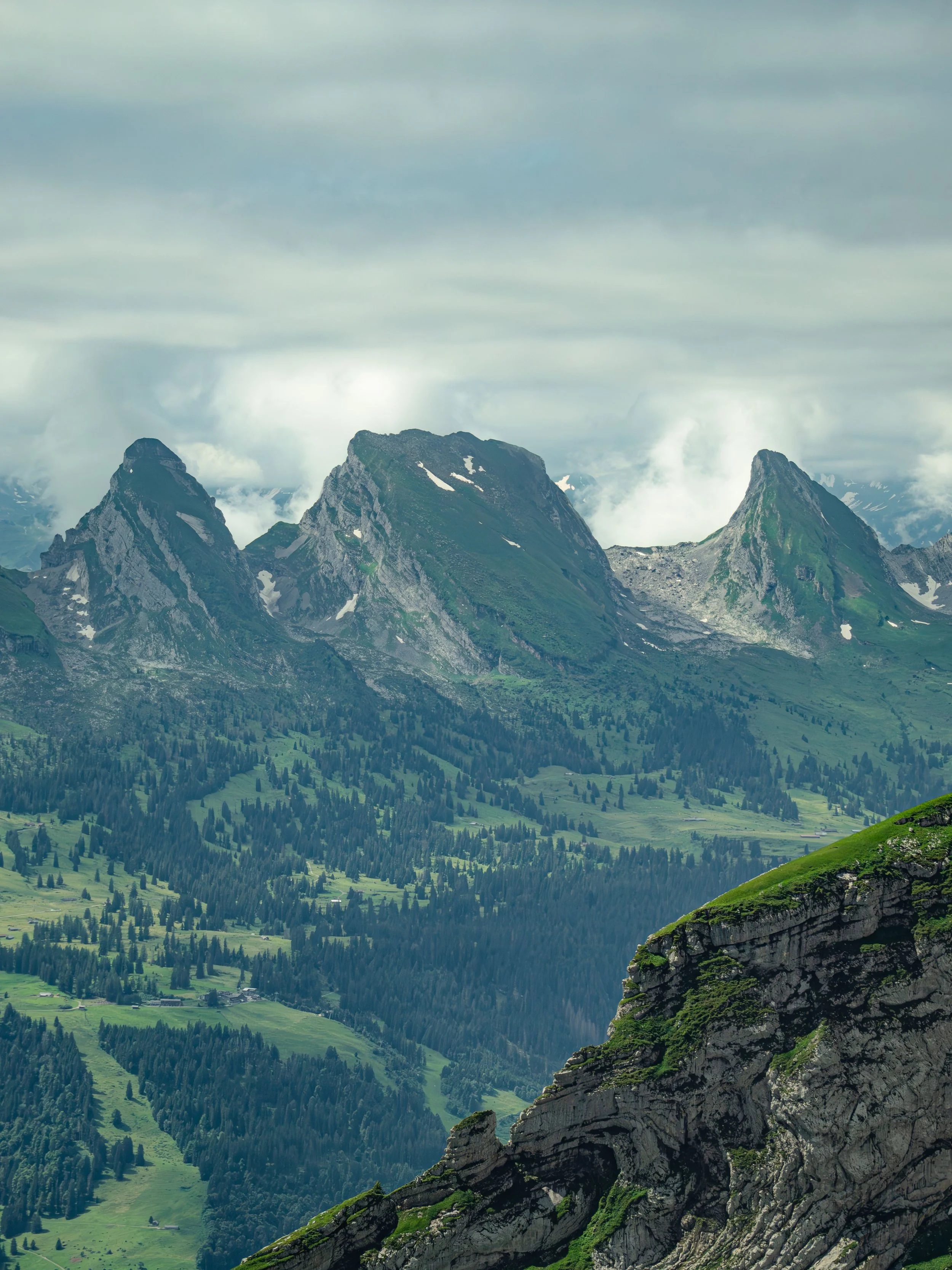 Montagnes verdoyantes avec des sommets rocheux, nuages et un ciel gris, vallées et forêts en contrebas.