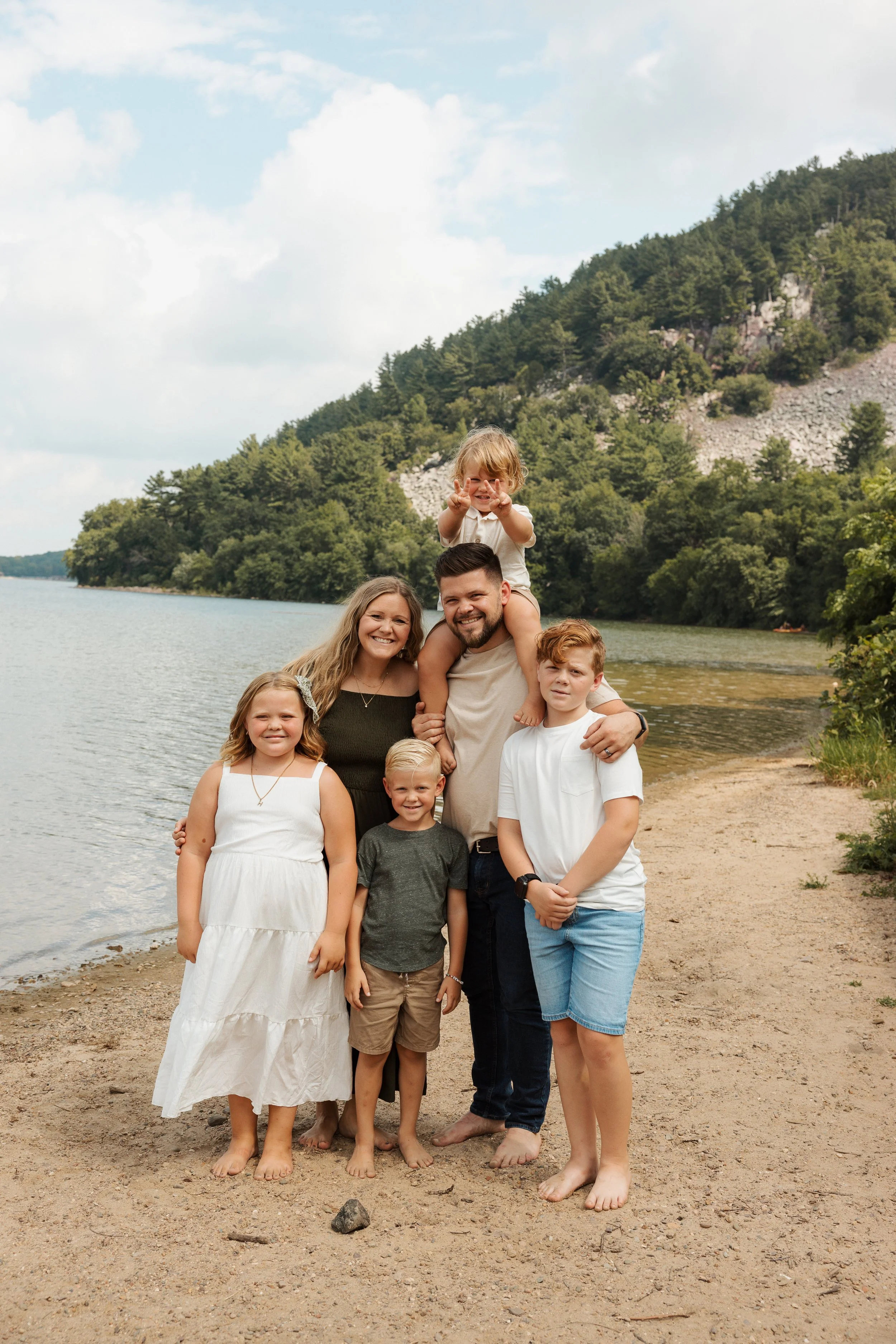 family of seven standing on a beach near a lake, with a forested hill in the background. The family includes two adult women, two adult men, and three children, one of whom is sitting on a man's shoulders.
