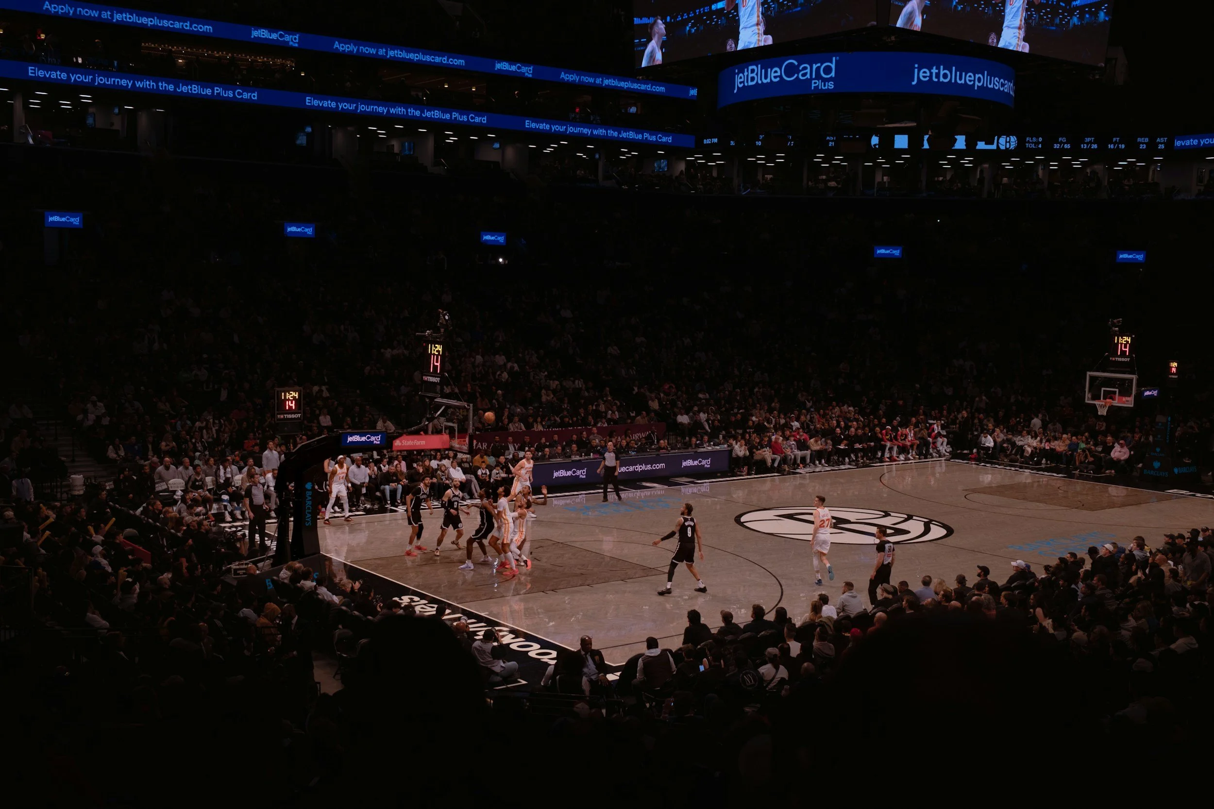 An indoor basketball game being played with players on the court and spectators in the stands. Large digital screens display advertisements for JetBlue and other sponsors.