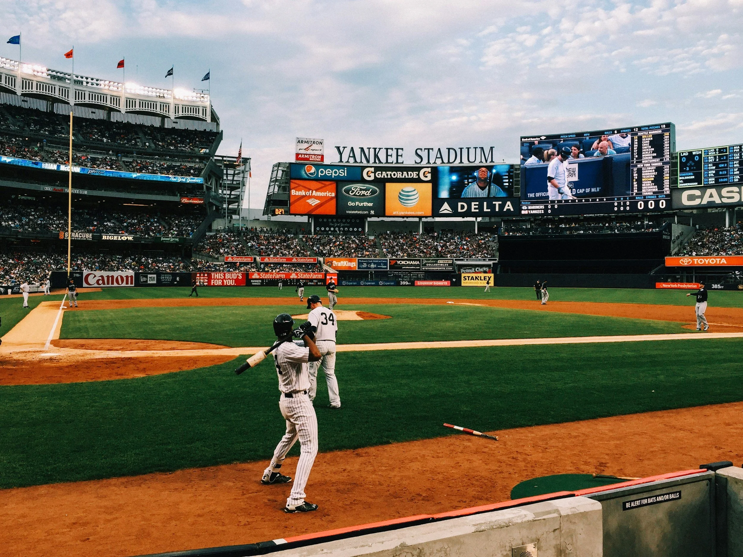 Baseball field at Yankee Stadium with players and a large video scoreboard showing team information, advertisements, and a close-up of a player. Fans are in the stands and the sky is partly cloudy.