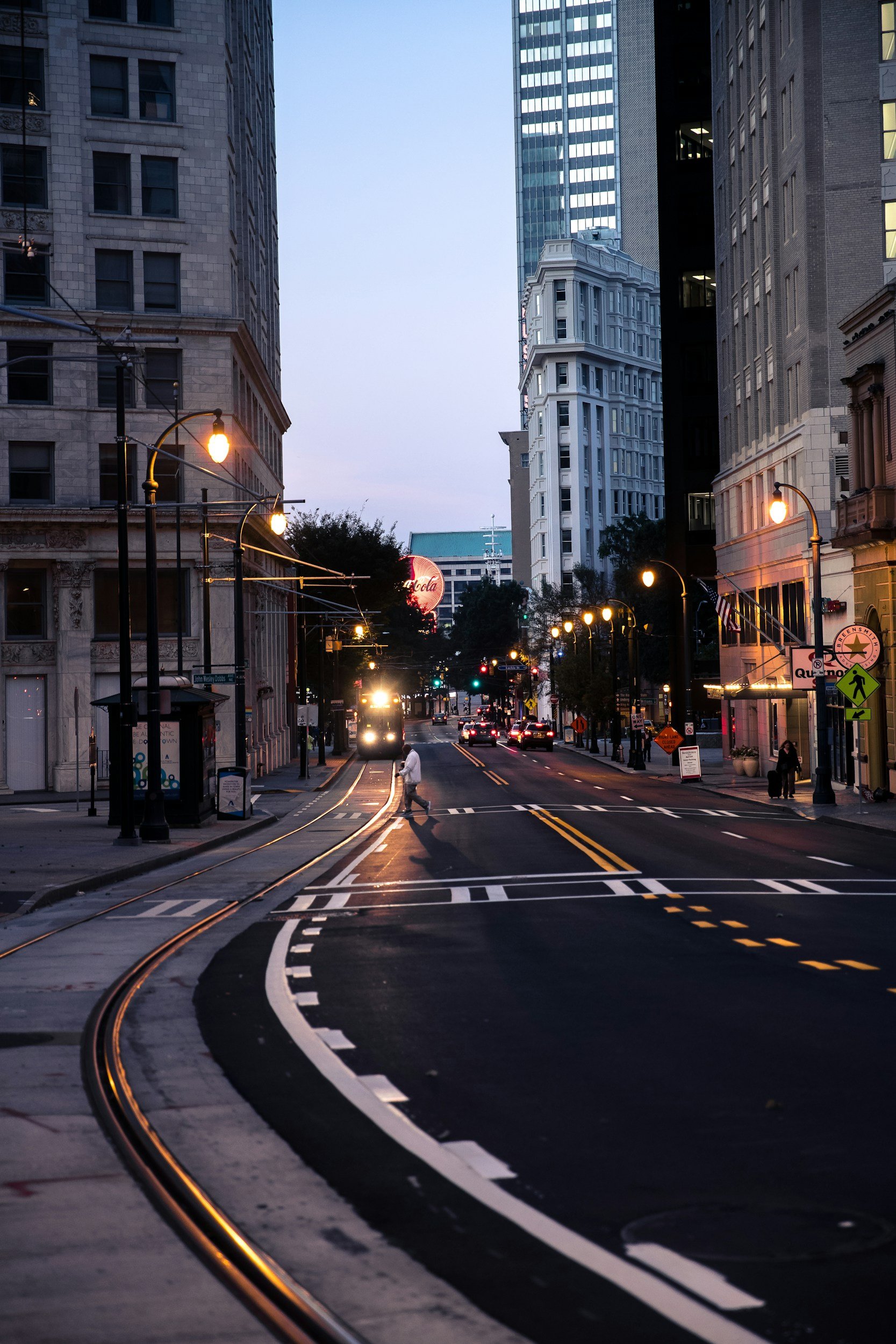 City street at dusk with tall buildings, traffic lights, and a streetcar track. A man with umbrella crossing the street. Intersection and street signs visible.