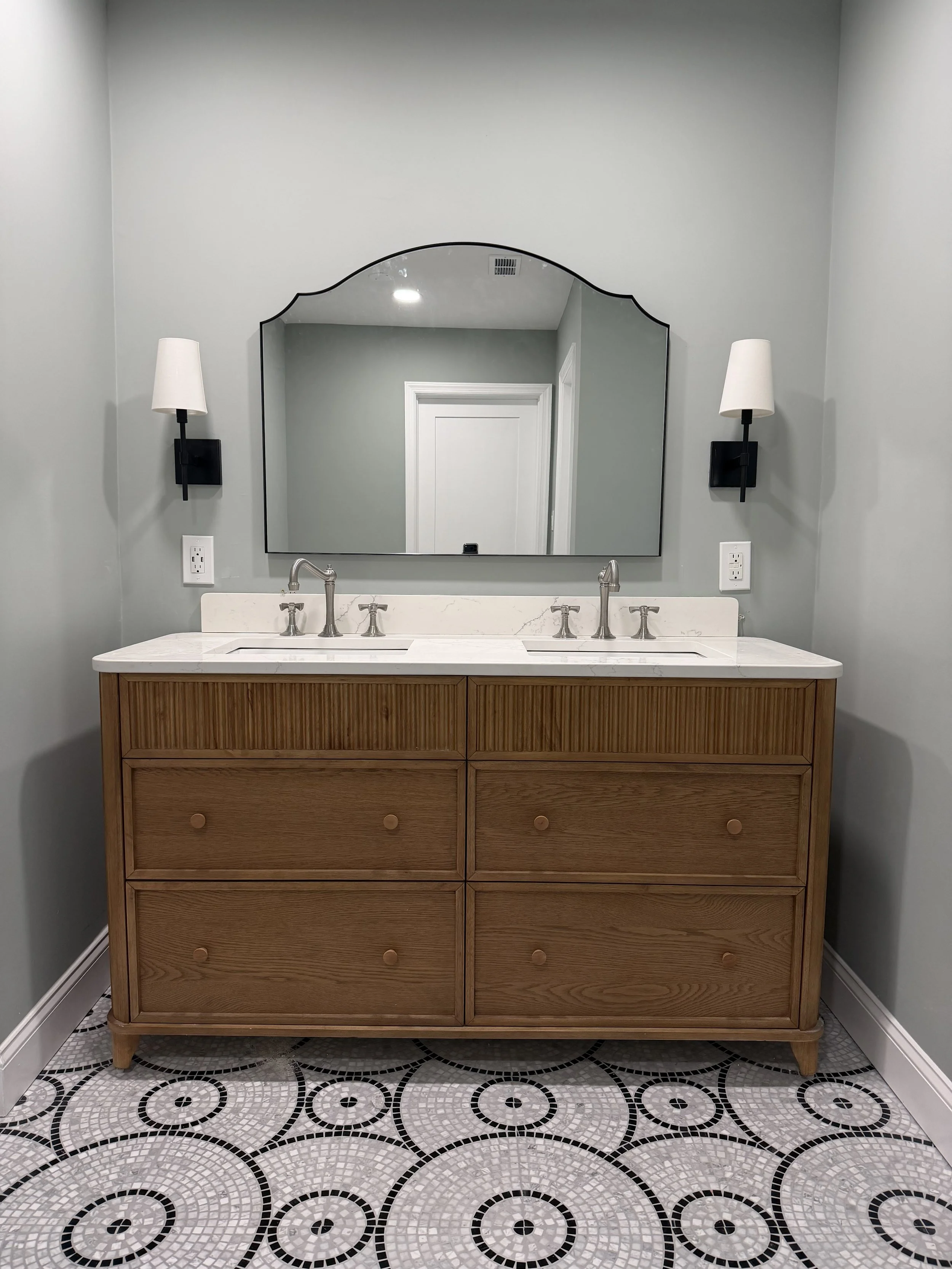 Bathroom vanity with a wooden cabinet, marble countertop, dual sinks, large mirror, wall-mounted sconces, and patterned tile floor.