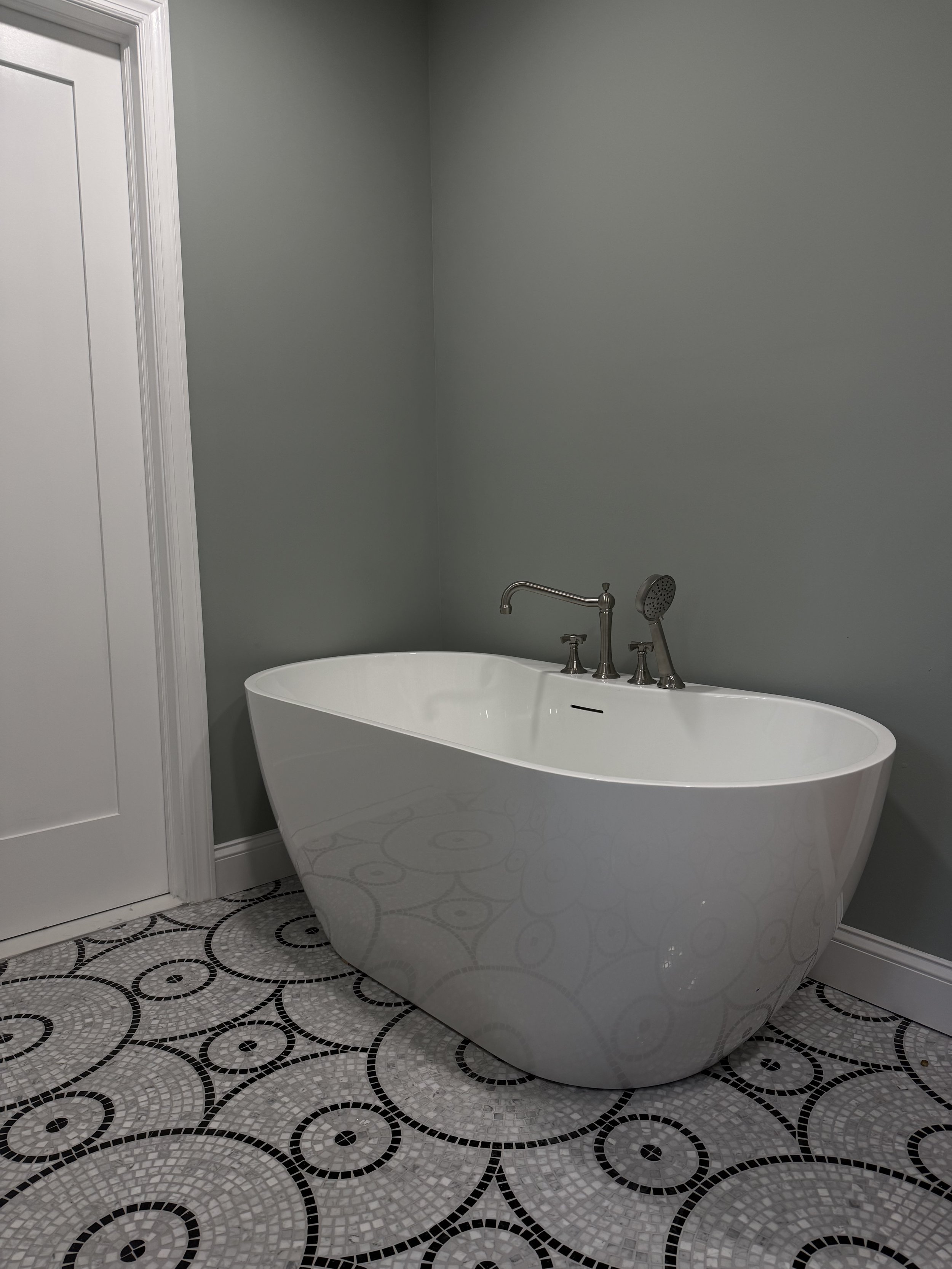 Freestanding white bathtub with a curved design, silver faucet, and handheld showerhead in a bathroom with gray walls and black and white patterned tiled floor.