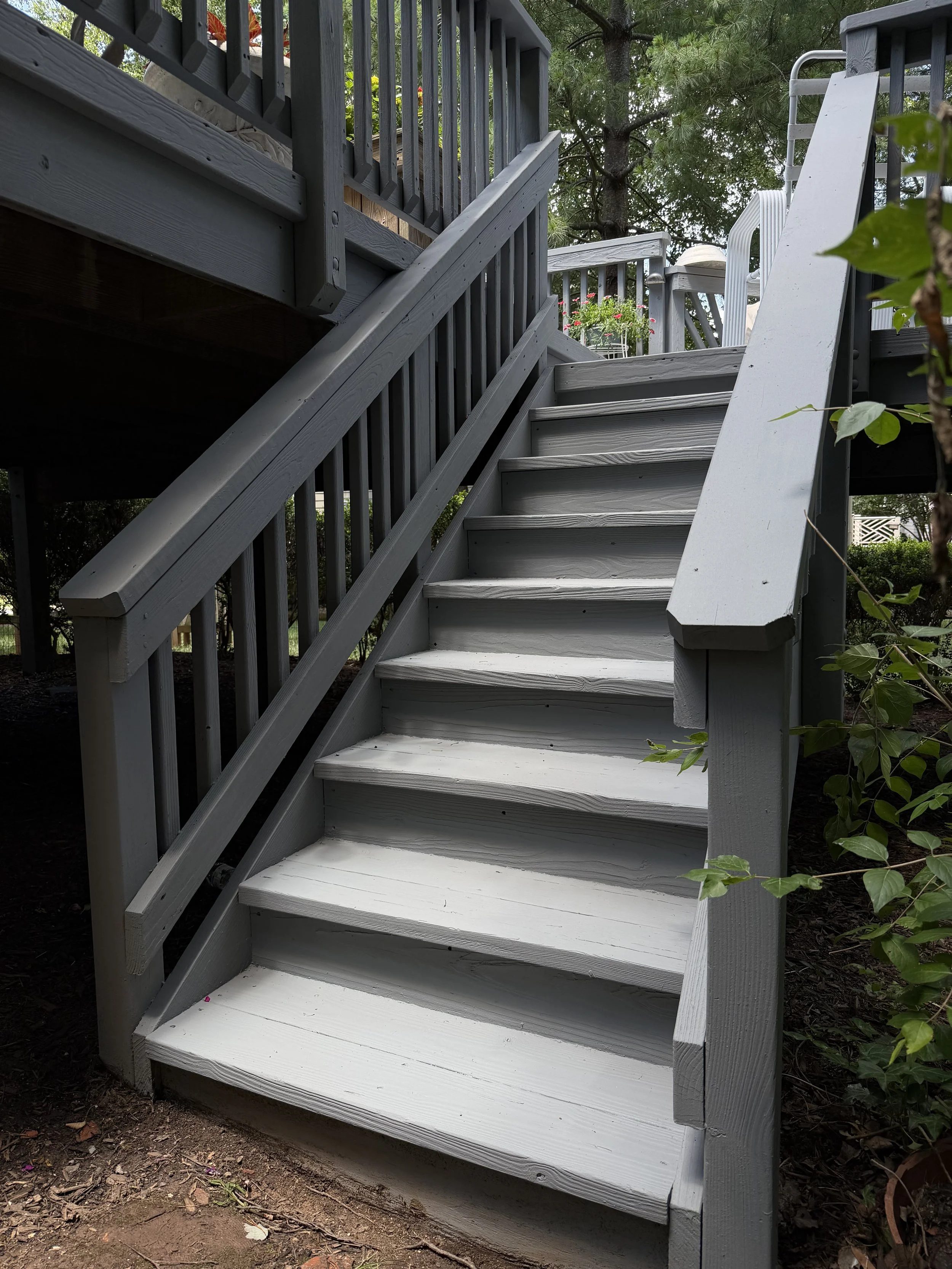 A staircase leading up to a deck with potted flowers and outdoor furniture, surrounded by trees and greenery.