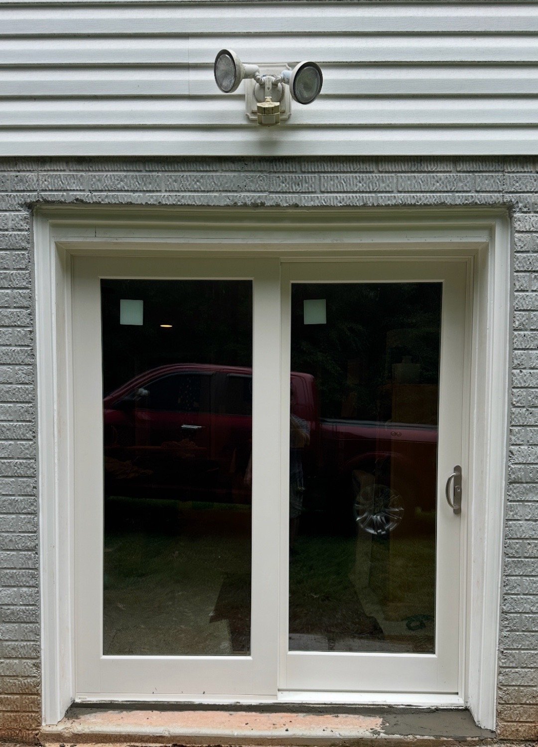 A white sliding glass door with a white frame, set in a brick and siding exterior wall, with a security light fixture above.