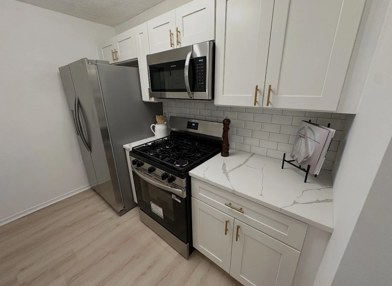 Modern kitchen with white cabinets, stainless steel refrigerator and oven, microwave, and a white marble countertop with subtle veining. A cookbook stand with a towel, a pepper mill, and a white pitcher with wooden spoons are on the counter.