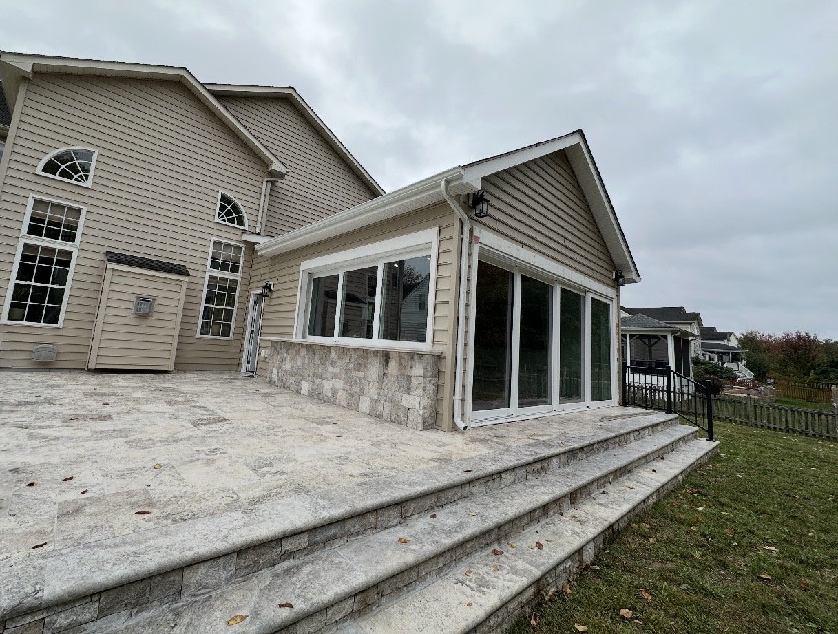 View of a modern residential house exterior showing a stone patio with stairs, beige siding, large windows, and a sunroom with sliding glass doors, under a cloudy sky.