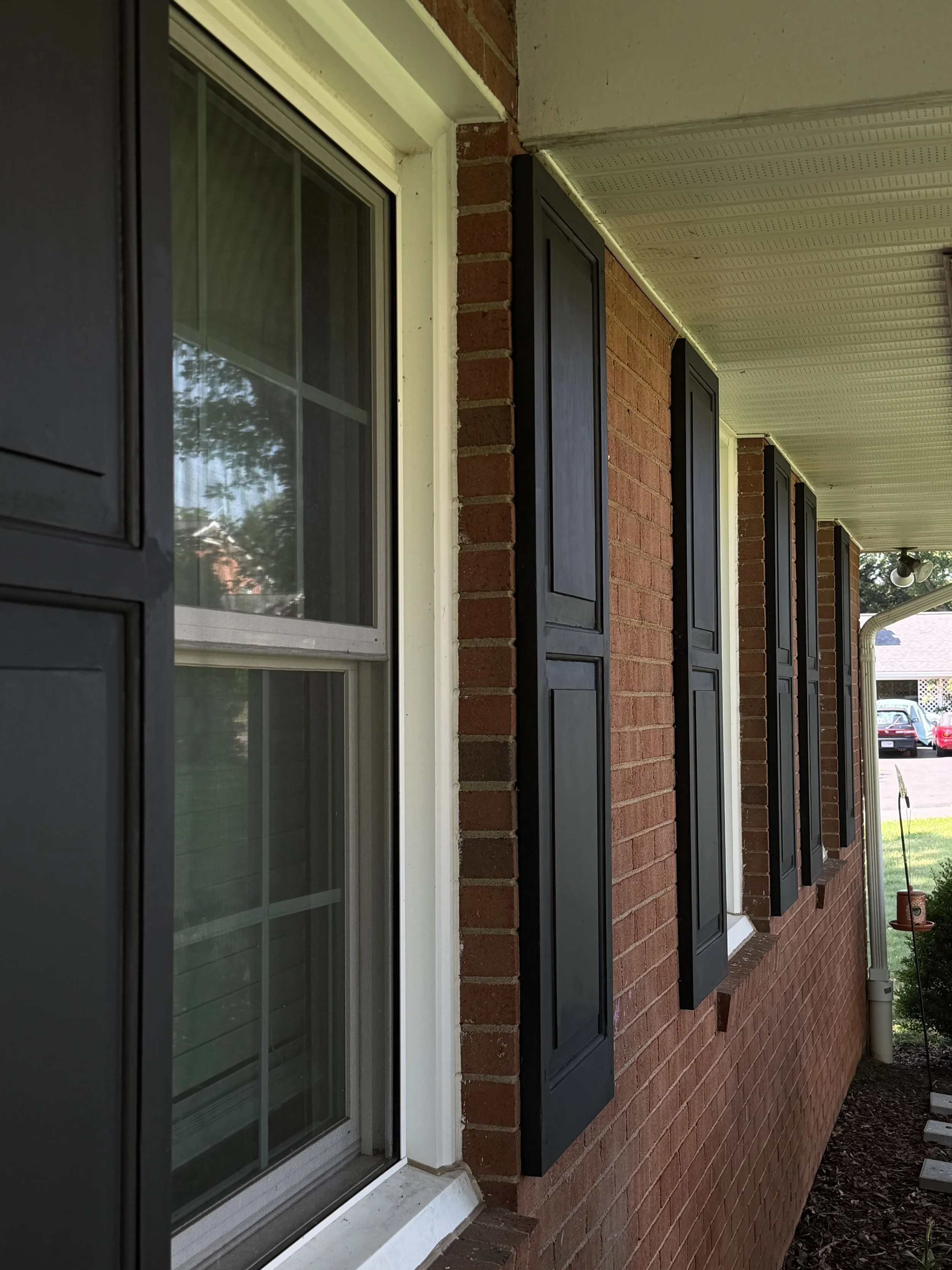 Close-up of a brick house exterior with a sliding glass door and four shuttered windows with black shutters, a white window frame, and black shutters, along with a drainage pipe visible in the yard.