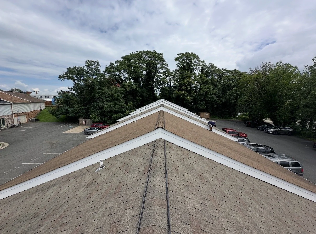 View from the top of a building's roof, showing a person working on the shingles with a parking lot and trees in the background.