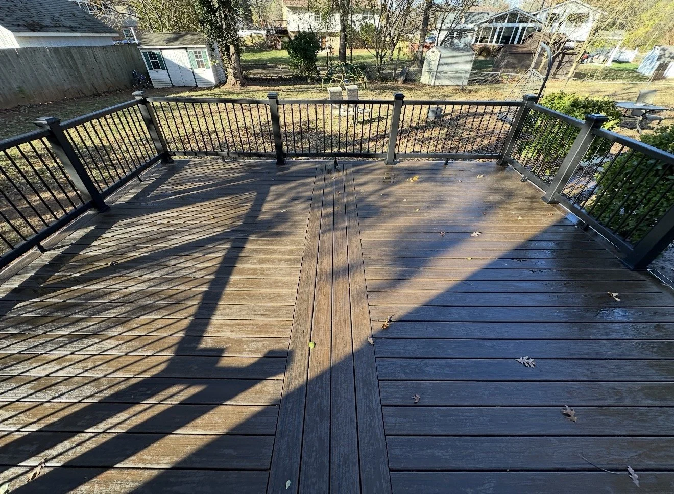 A wooden backyard deck with black metal railing, cast shadows, and some fallen leaves. In the background, there are trees, sheds, and a greenhouse.