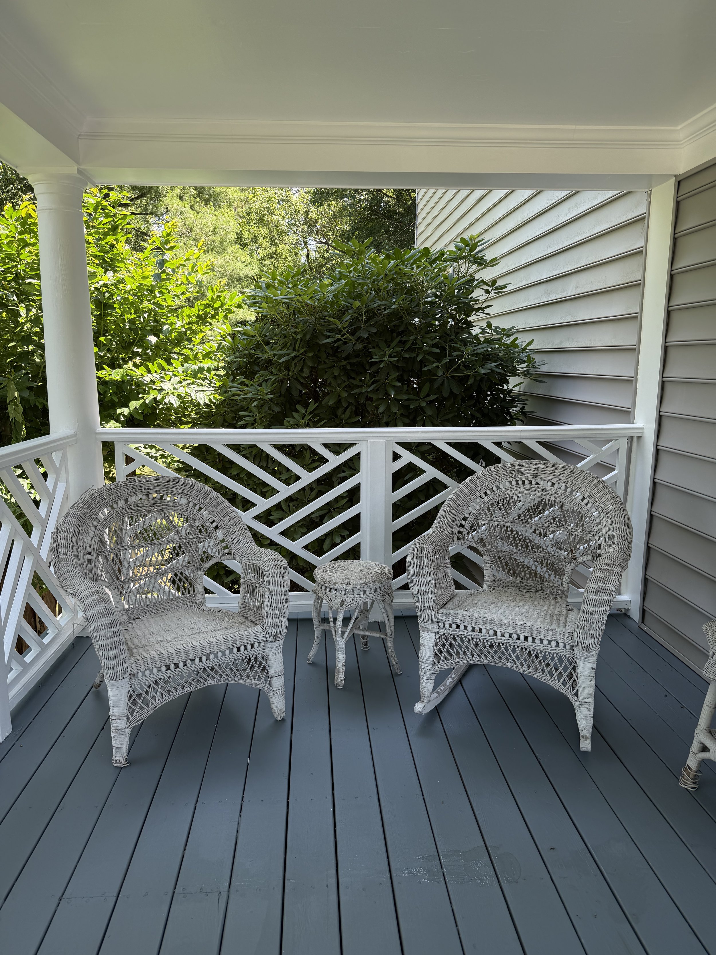Porch with a white lattice railing, three white wicker chairs, a matching round table, green bushes and trees in the background, and gray wooden floorboards.