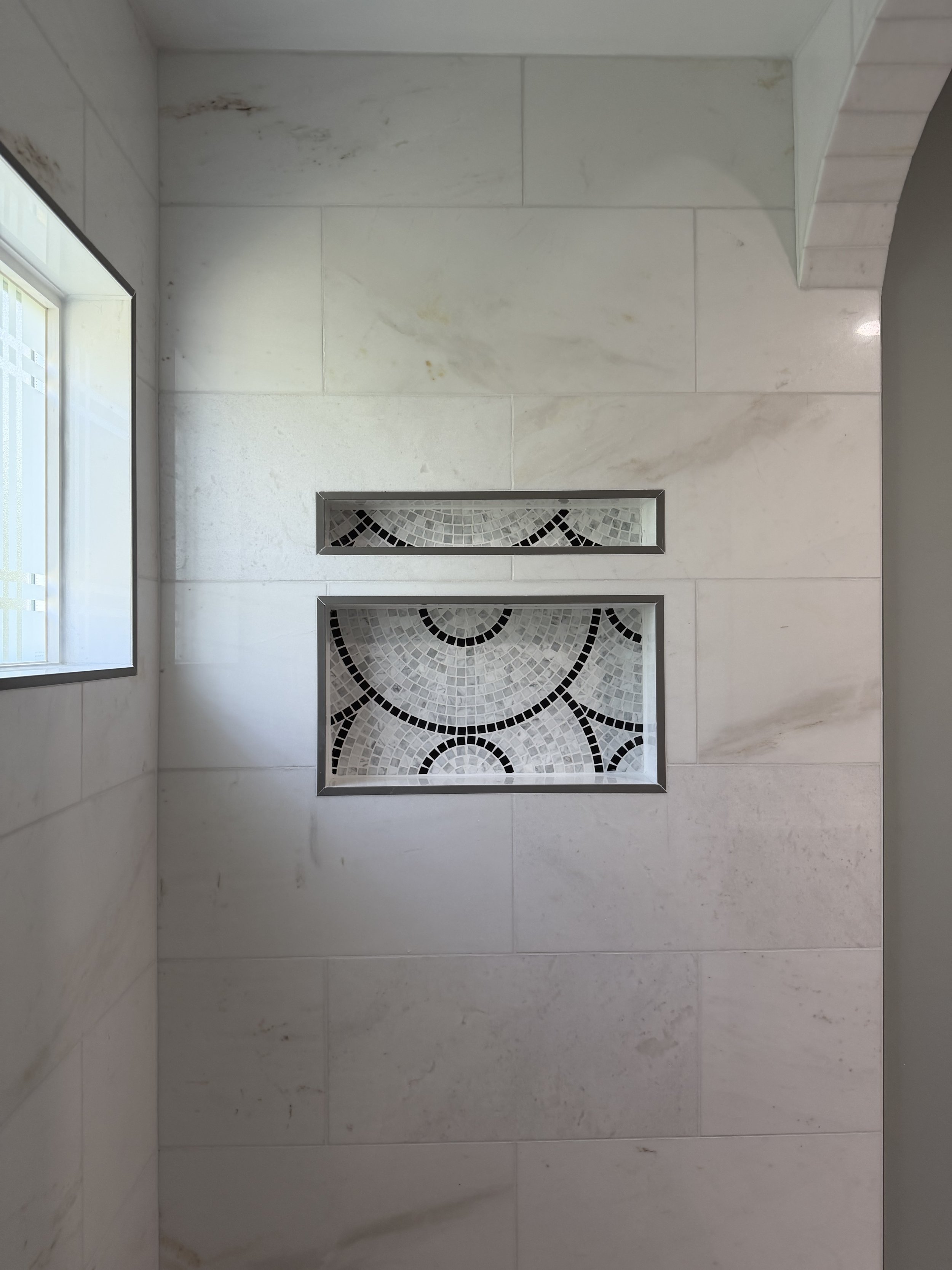 Bathroom wall with white marble tiles, two rectangular decorative insets with black and white mosaic patterns, and a window on the left side.