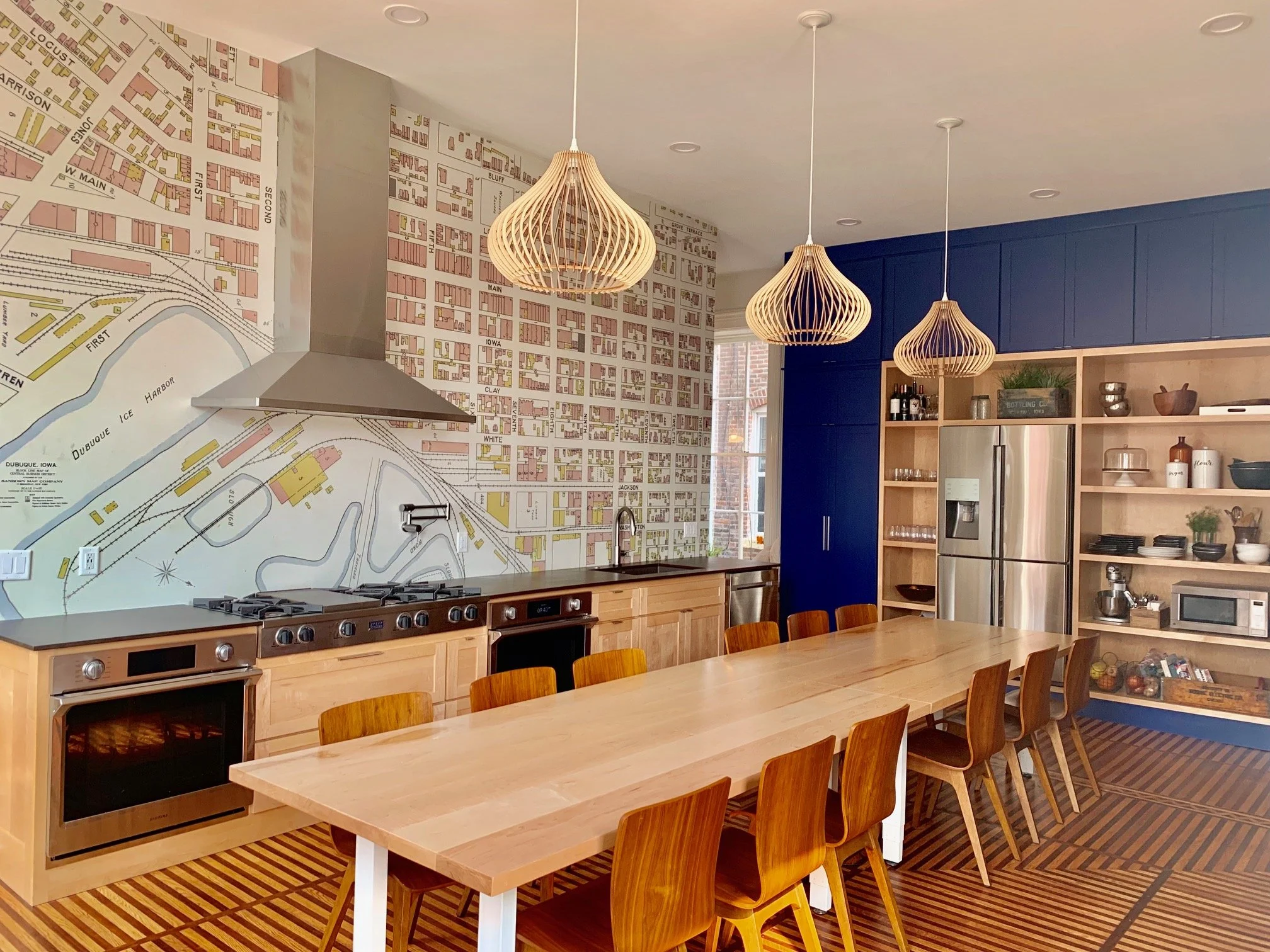 A kitchen with a long wooden dining table and chairs, blue cabinets, a stainless steel refrigerator, open shelving with dishes and plants, and a map mural on one wall illuminated by three wooden pendant lights.