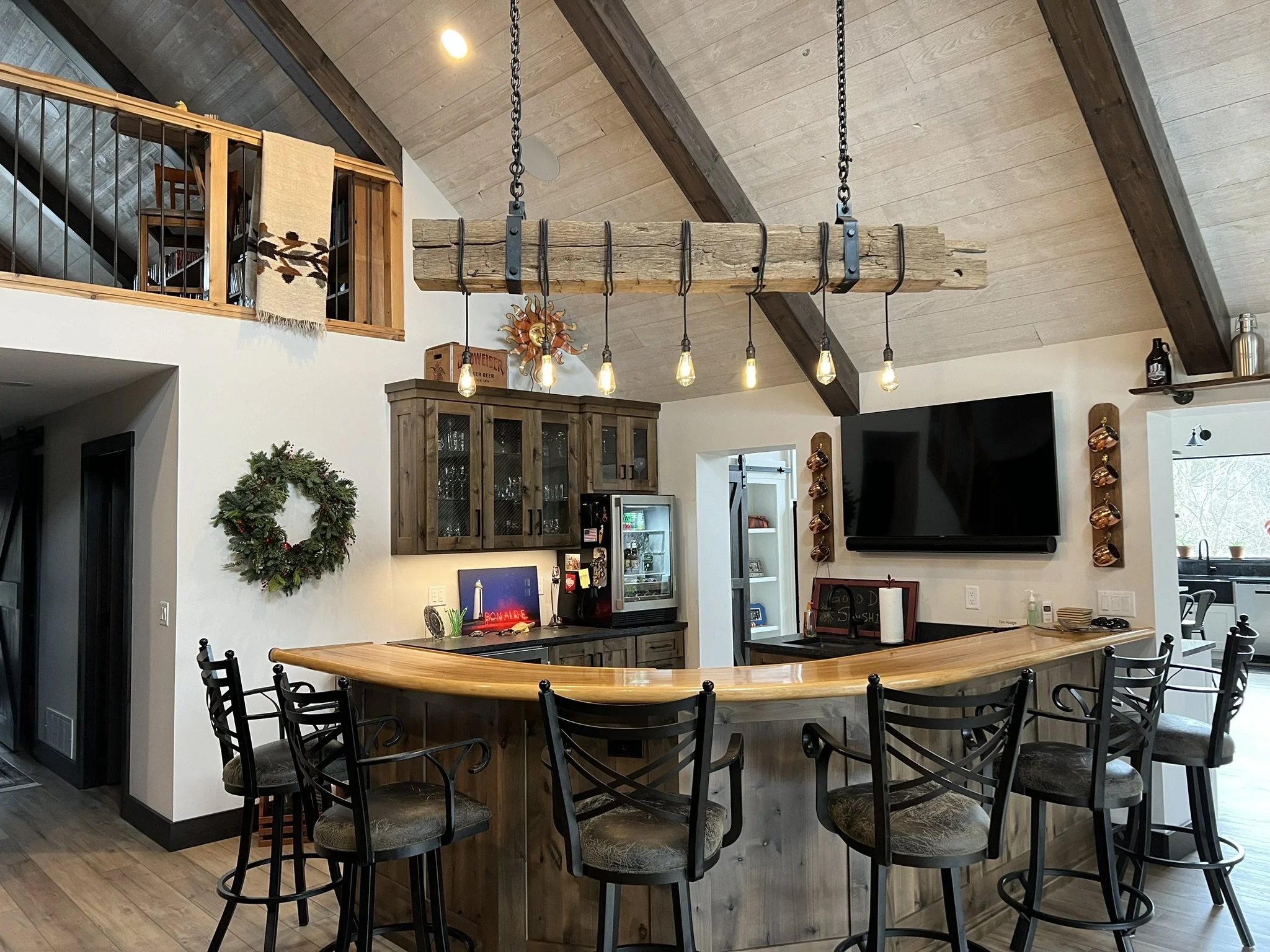 Interior view of a kitchen with a wooden island and black chairs around it, a wooden cabinet, a wall-mounted TV, and a rustic lighting fixture with Edison bulbs hanging from chains.