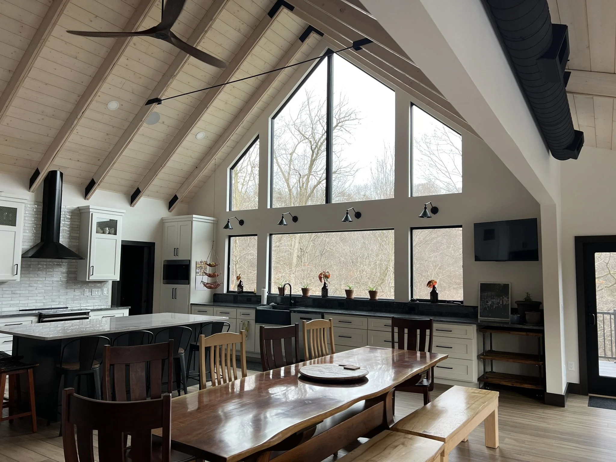 Open-concept kitchen and dining area with large triangular window, white cabinets, black countertops, and a high vaulted ceiling with exposed wood beams and a ceiling fan.