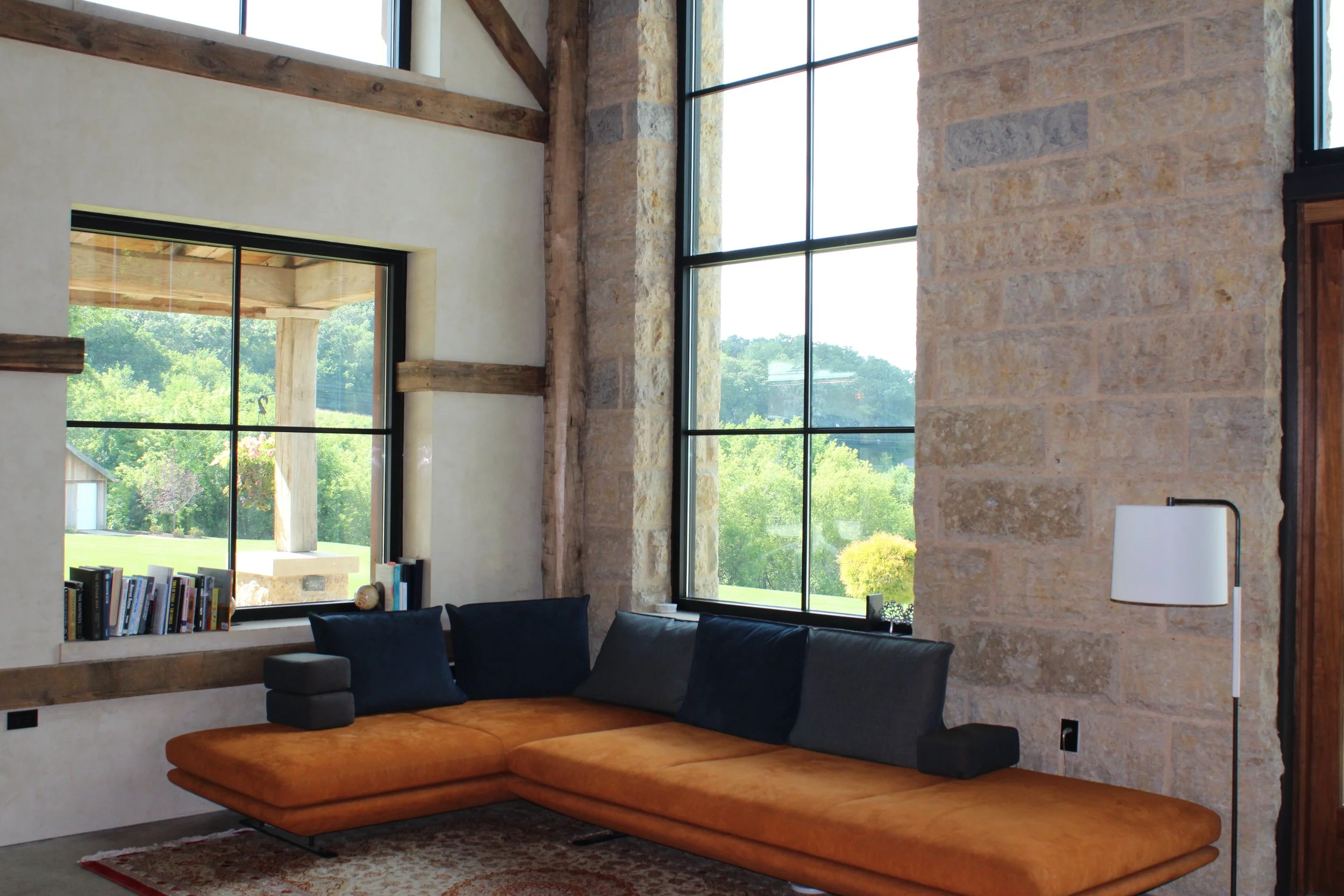 Living room with large windows, stone walls, wooden beams, a bookshelf, black sofa with orange cushion, and a white floor lamp.